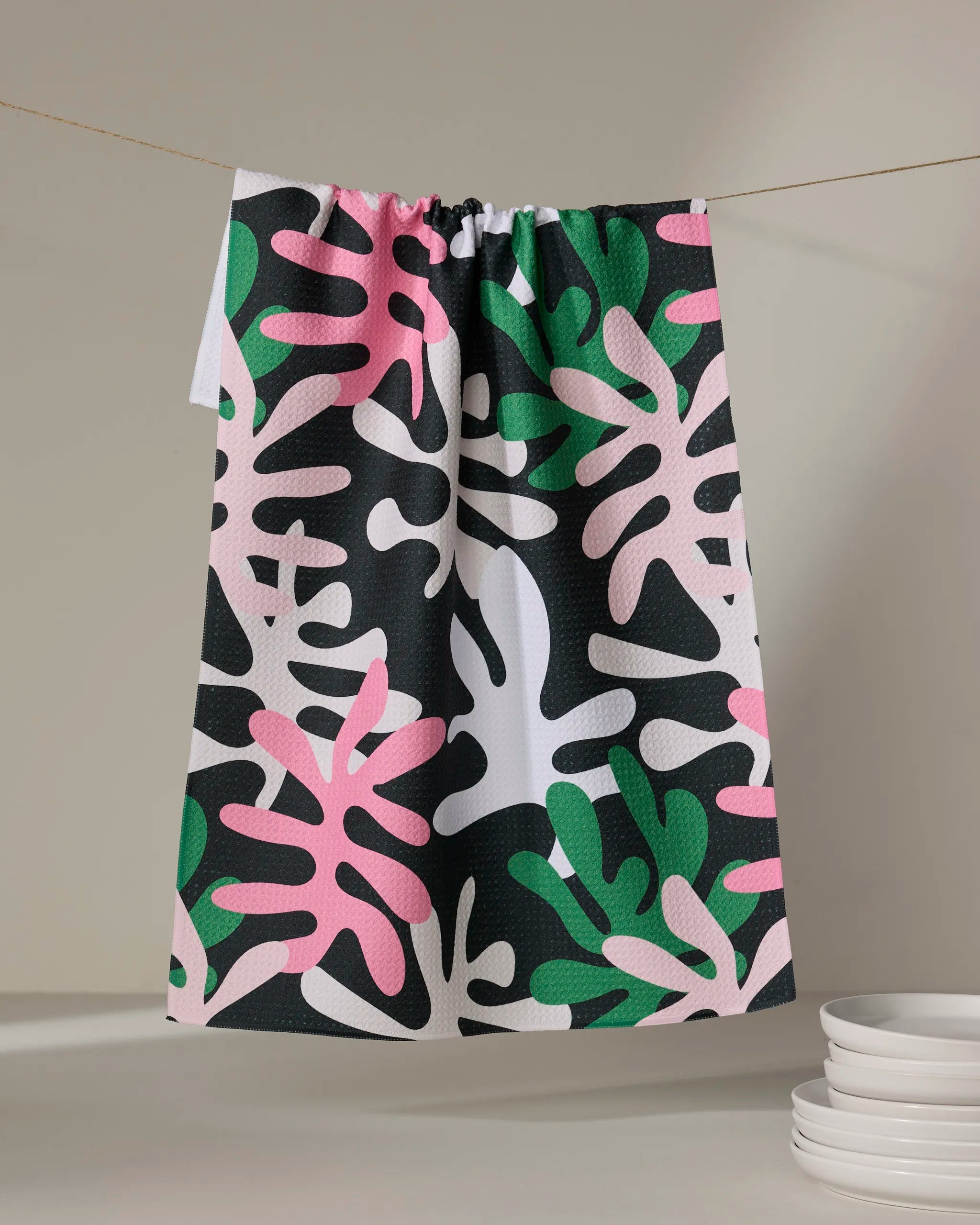 The Late Night tea towel by Marina Solodka features a bold abstract leaf pattern in pink, green, black, and white. Made from recycled materials, it adds style near stacked white plates on a neutral surface.