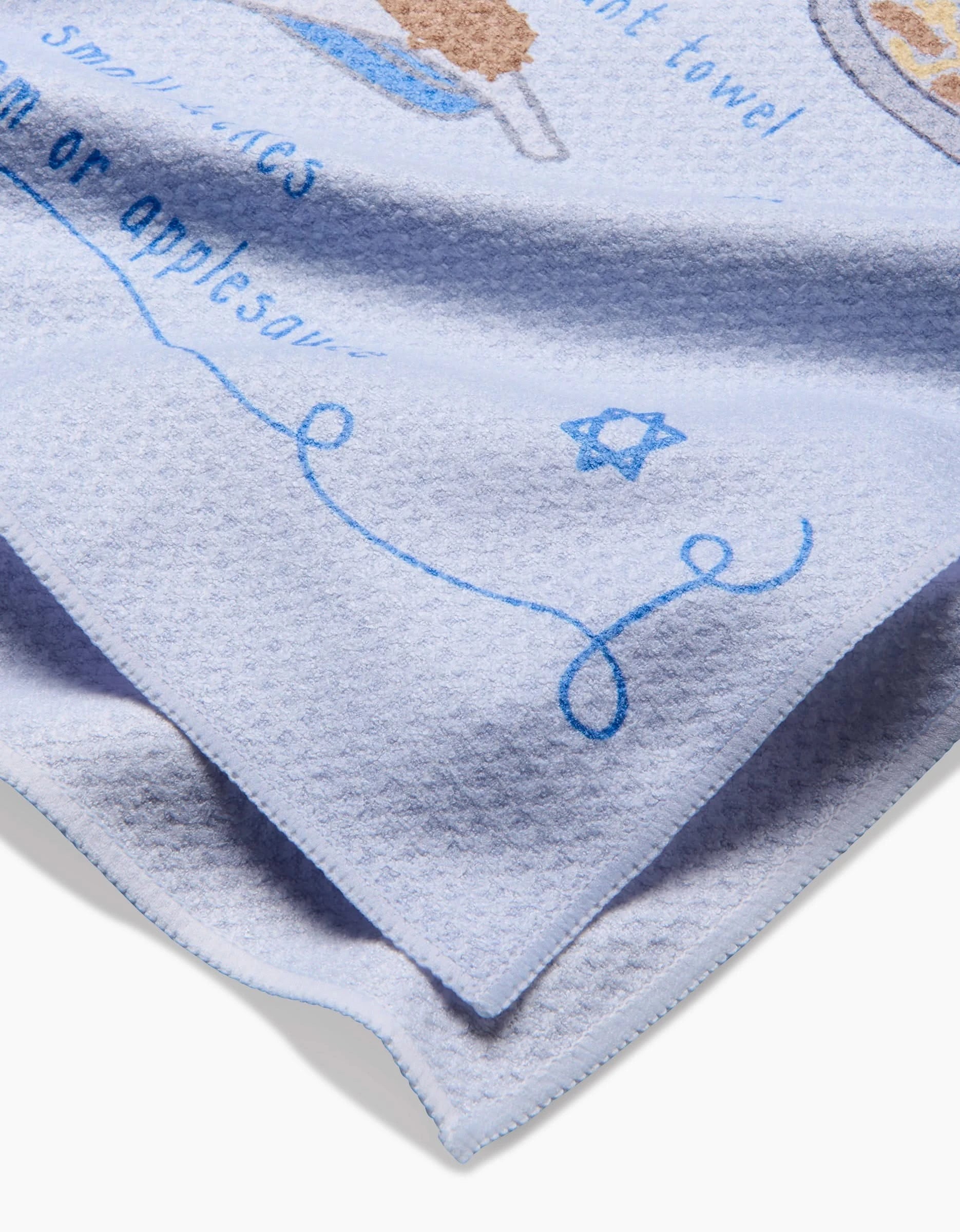 A close-up of the Latke Love tea towel by Mindfulnice, featuring textured light blue fabric with blue text, a Star of David, part of a line drawing, and printed food graphics on this quick-drying dish towel.