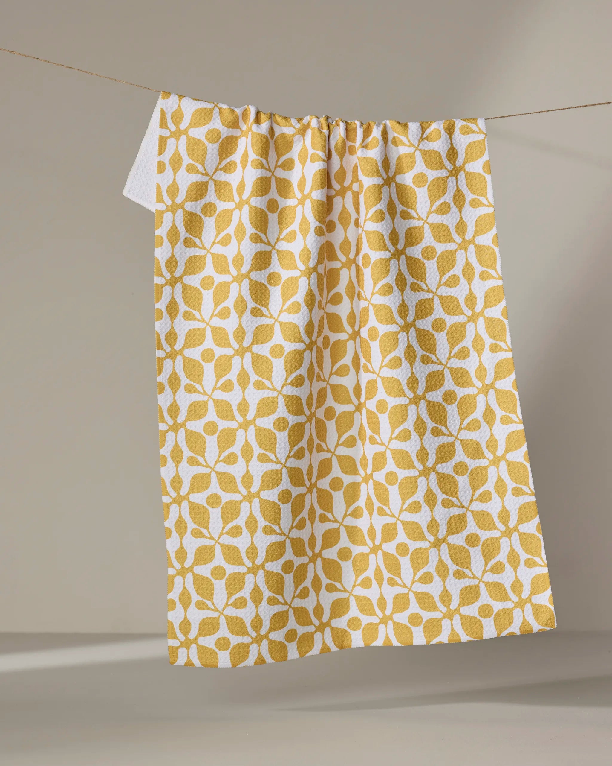 The Renee Fly "Lattice" dish towel, featuring a yellow and white geometric floral pattern, hangs on a clothesline against a neutral beige background.