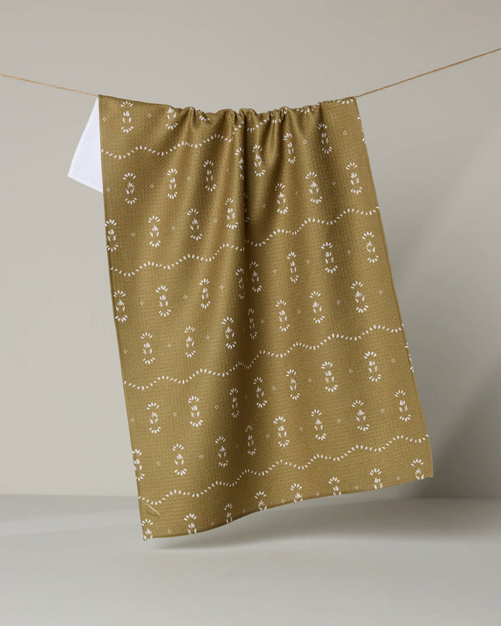 The Laurel tea towel by Melissa Johnson is brown with a white floral and dotted pattern, made from recycled materials. It hangs on a clothesline against a light gray background.
