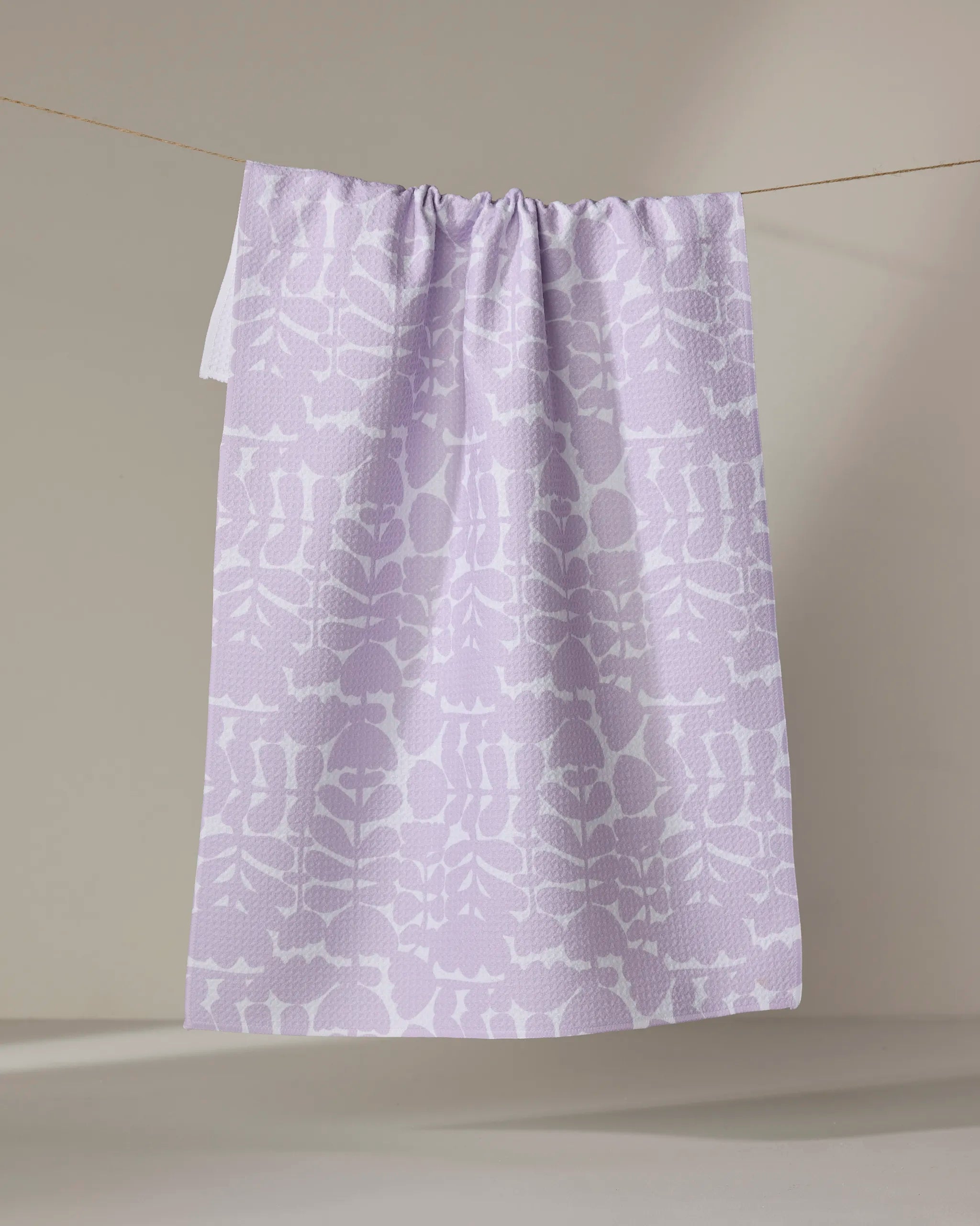 The Katie O'Shea Lavender Scandi Flowers tea towel, featuring a white botanical pattern and made from quick-drying recycled materials, hangs on a thin clothesline against a neutral beige background.