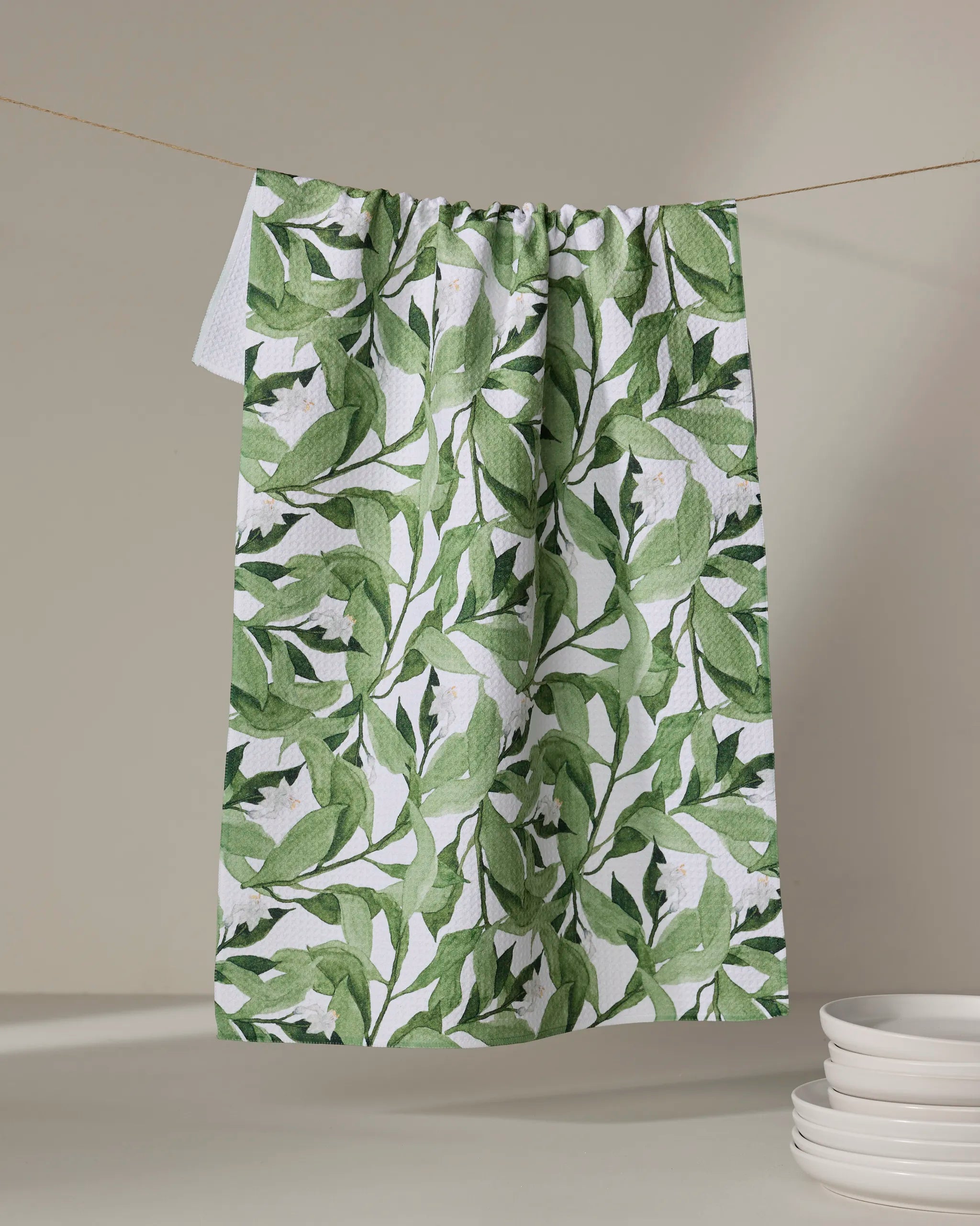 The Aubrey Fairchild Leaf It To Me dish towel, featuring a green and white leafy botanical pattern, hangs from a clothesline above stacked white plates on a neutral background.