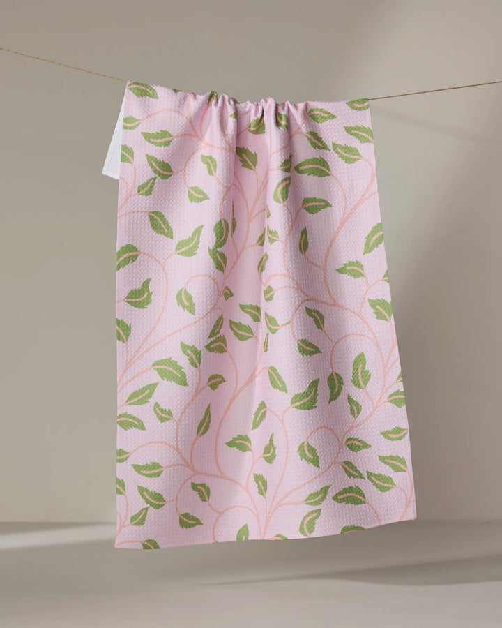 The Leafy Maze tea towel by Jasmine Blooms Designs features a pink fabric with green leaf patterns, made from recycled materials. Its subtly textured cloth is gathered at the top and hangs from a string against a neutral backdrop.