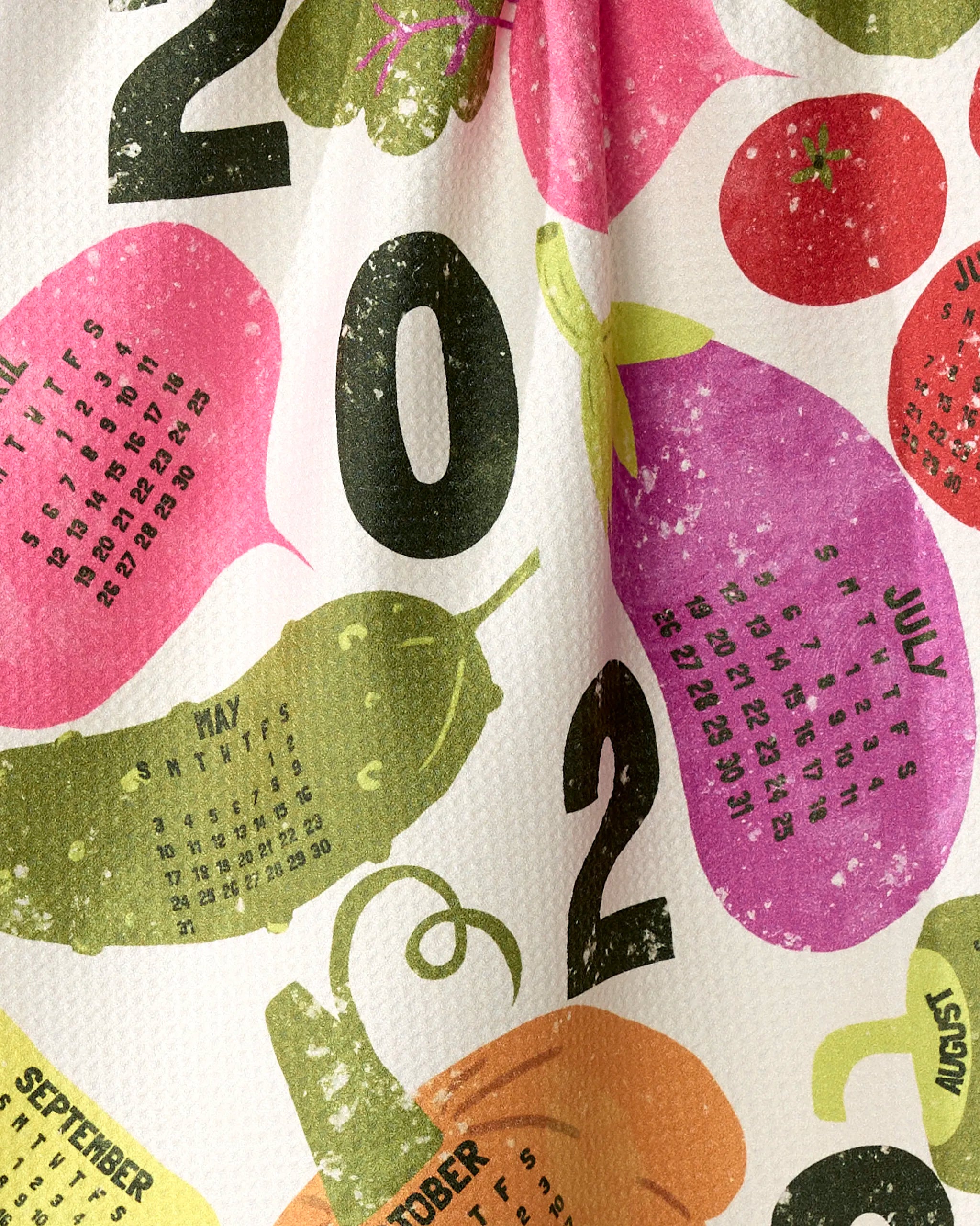 The Legume Calendar by Geometry is a vibrant tea towel made from recycled materials, featuring 2023 months printed among illustrated veggies and fruits like peppers, eggplants, and tomatoes, with bold black "2023" partially visible.