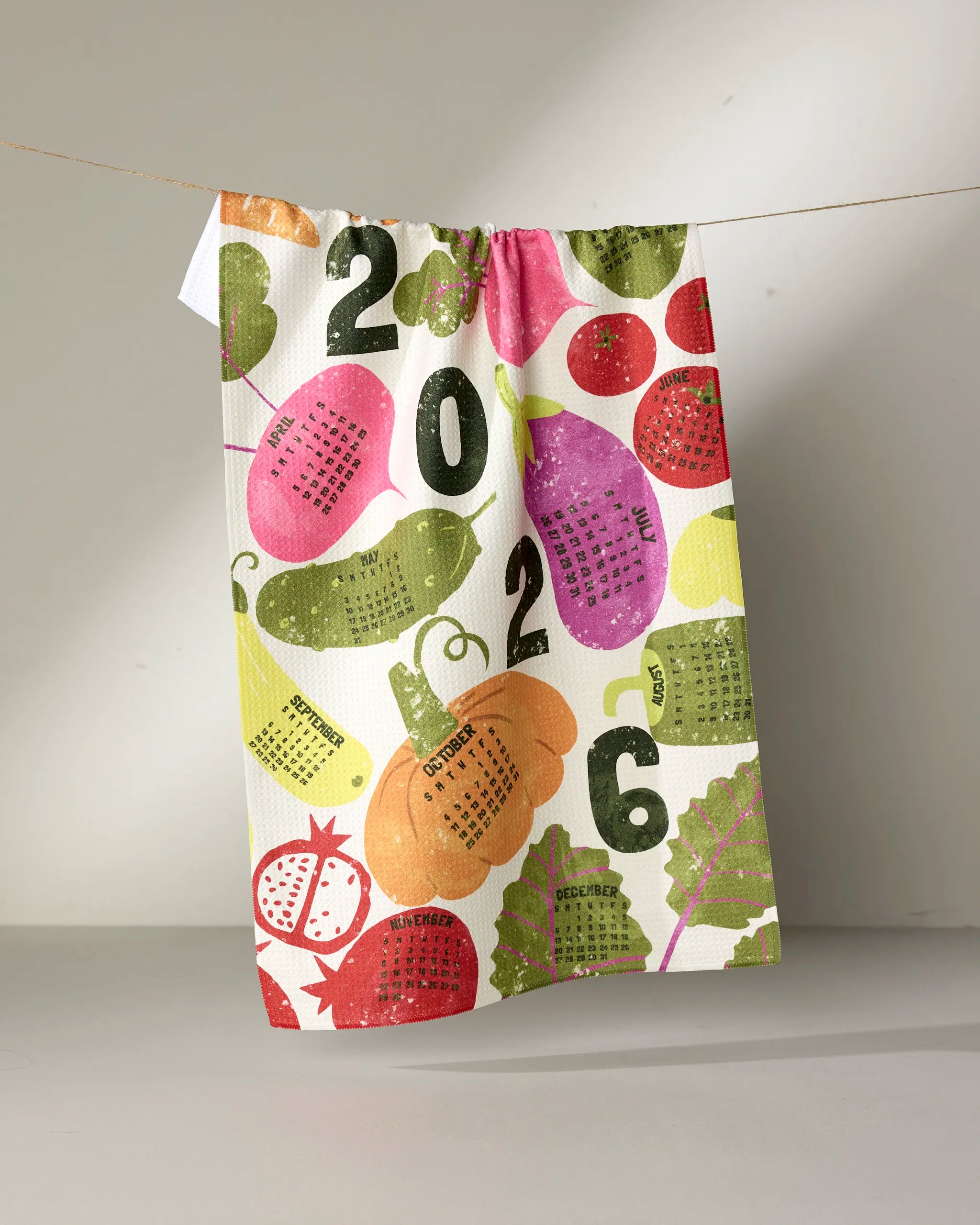 The Geometry Legume Calendar tea towel, made from recycled materials, displays each 2024 month inside vibrant illustrated fruits and vegetables. Hanging on a string, it brightens your kitchen with color and eco-friendly style.