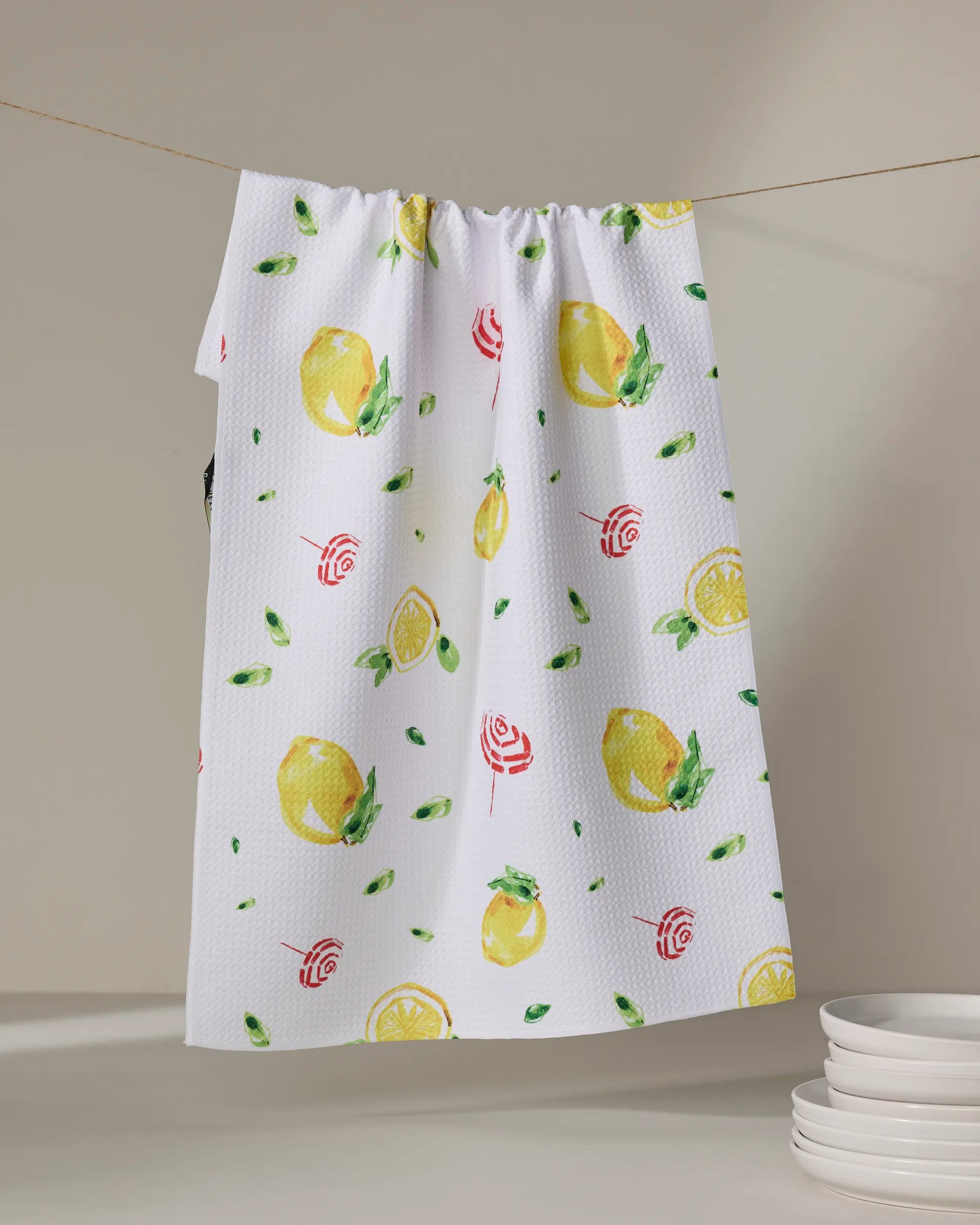 Lemons in Capri" by Travel Write Draw features a white tea towel with lemons, leaves, and red swirls—made from recycled materials—hanging above stacked plates against a neutral background.