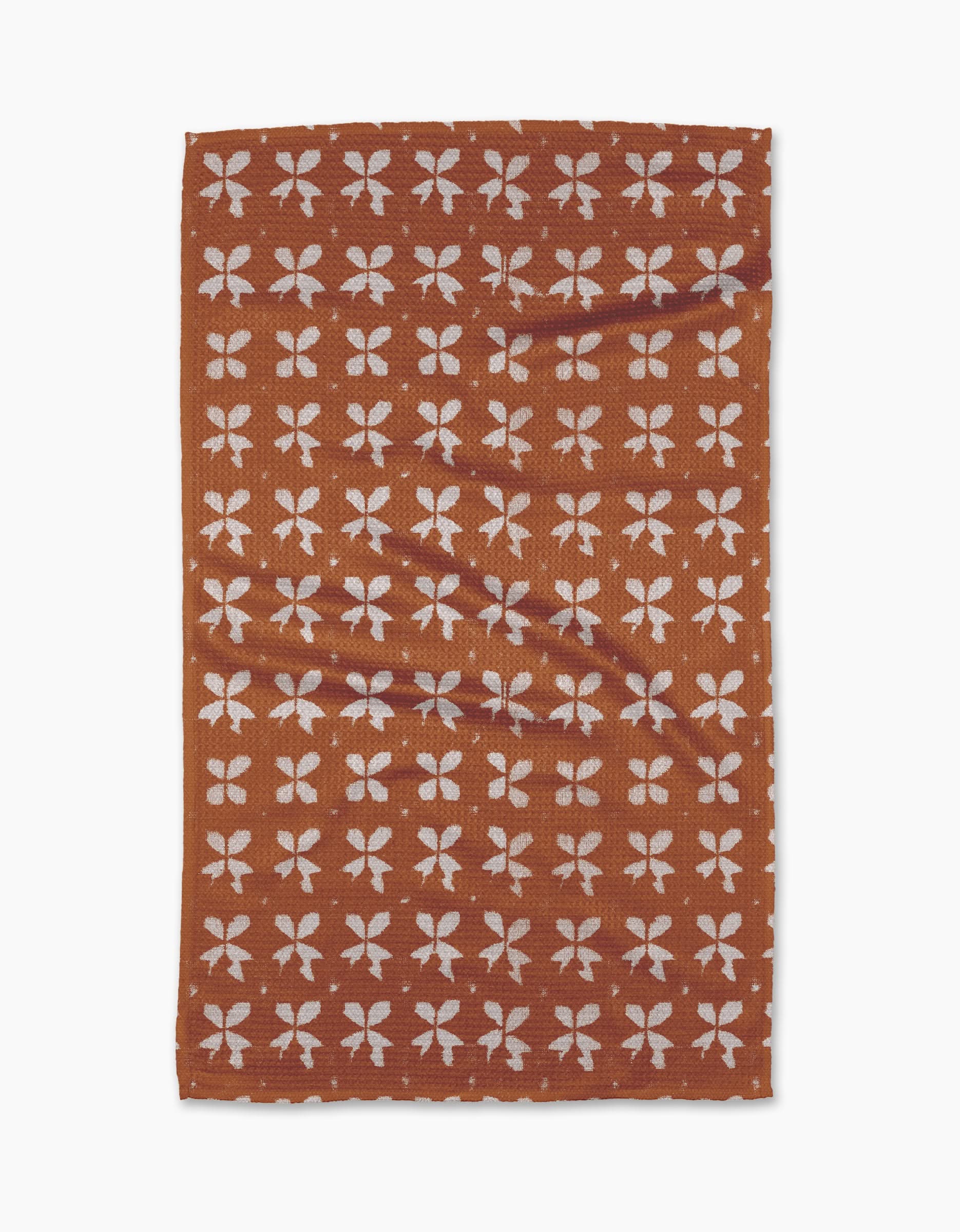 The Leonie tea towel by Geometry is rectangular and brown with a repeating pattern of white four-petaled floral shapes in rows. Made from eco-friendly fabric, it appears slightly wrinkled and is shown on a plain white background.