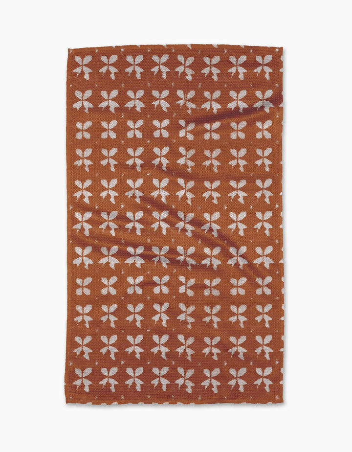 The Leonie tea towel by Geometry is rectangular and brown with a repeating pattern of white four-petaled floral shapes in rows. Made from eco-friendly fabric, it appears slightly wrinkled and is shown on a plain white background.