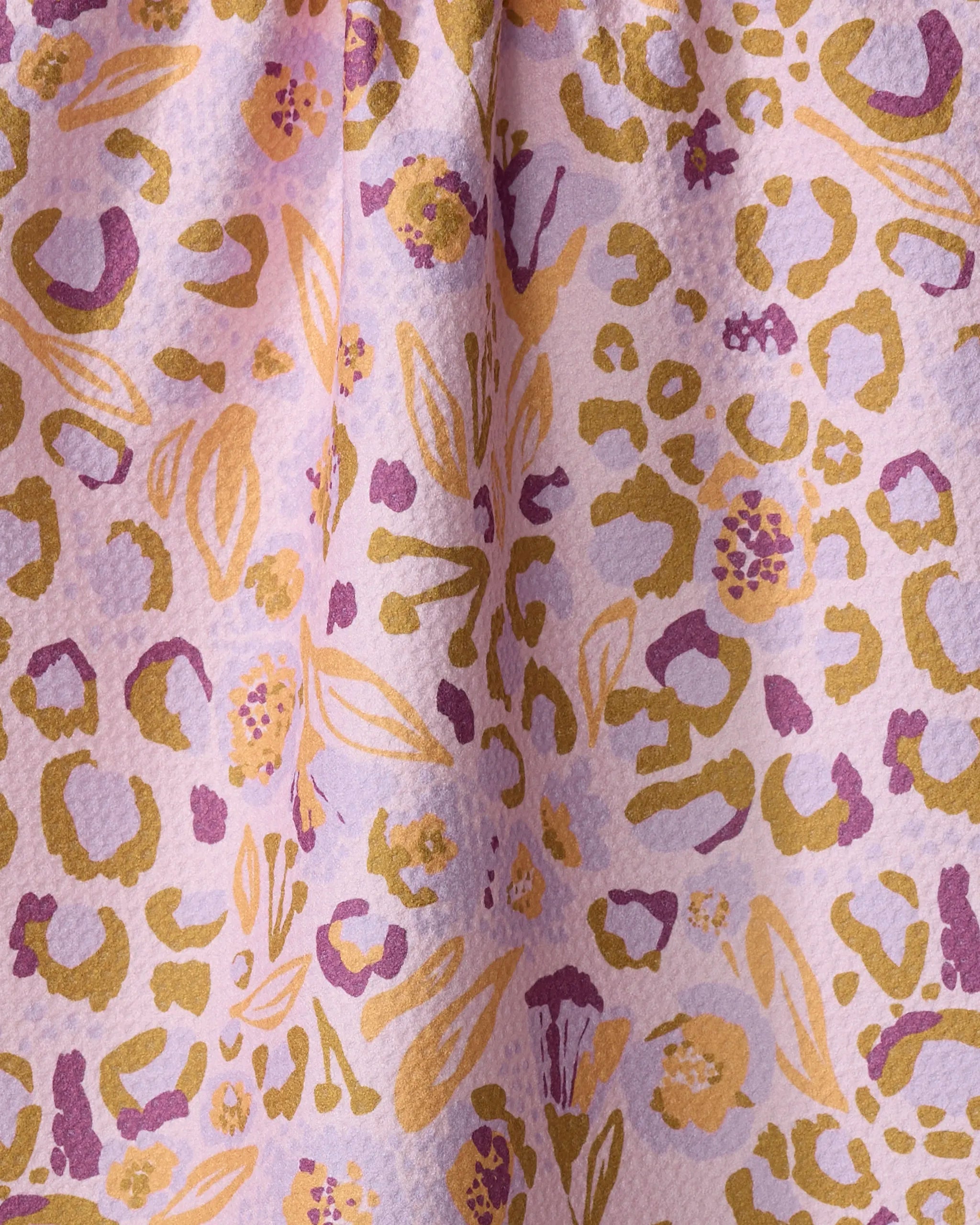 A close-up of Elisabeth Wing’s Leopard Loving kitchen towel, made from recycled materials, showcases a textured textile with gold, yellow, and purple abstract shapes and animal print spots on a pale pink background.