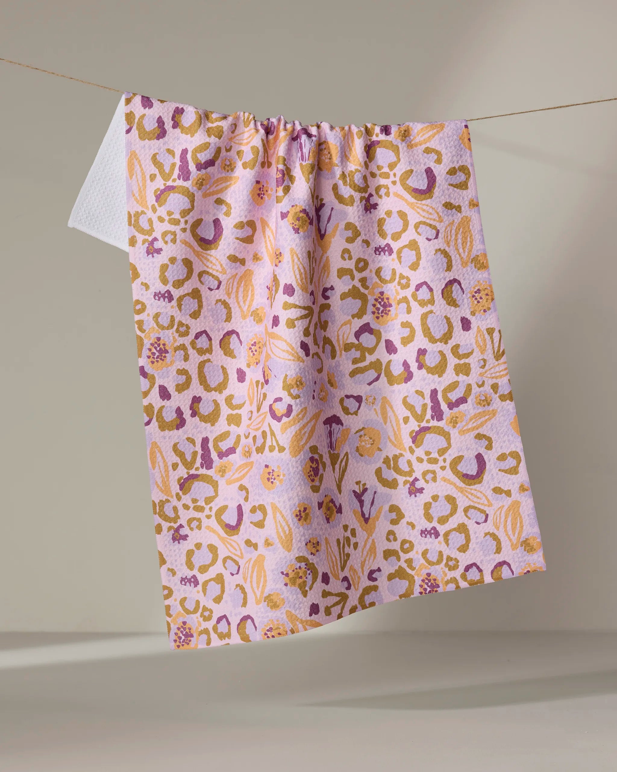 The Leopard Loving tea towel by Elisabeth Wing features a vibrant pink, orange, and purple leopard print and is made from recycled materials. It hangs from a string against a neutral background.
