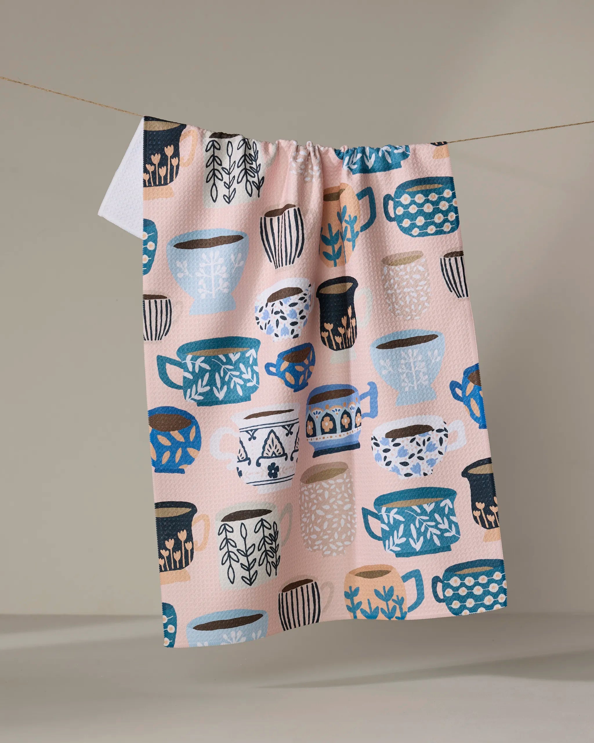 The Les Tasses tea towel by Cottonflower Studio features a vibrant pattern of mugs and cups in blue, black, white, and peach on recycled fabric, shown hanging on a clothesline against a neutral backdrop.