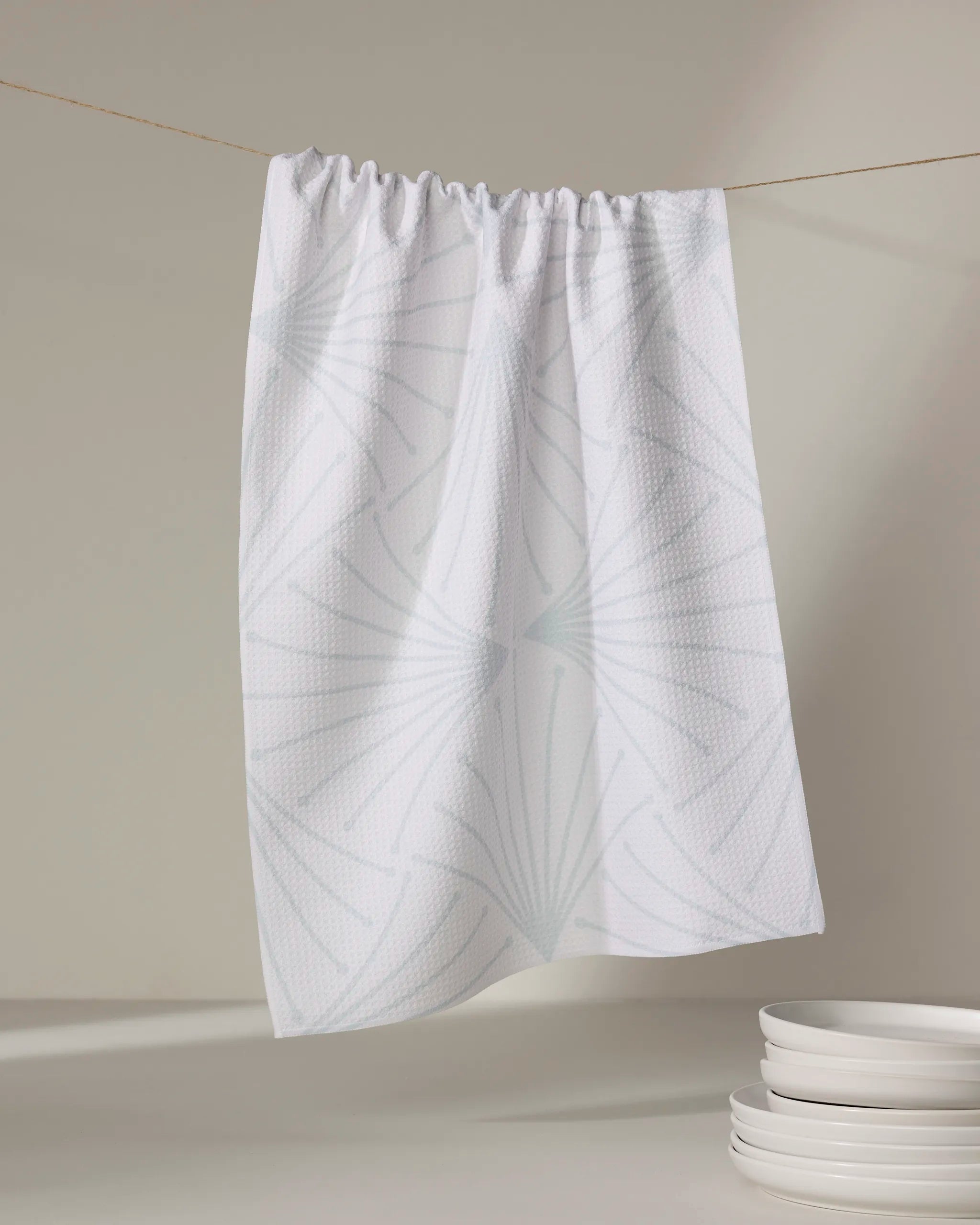 The Amy MacCready Libby Cream tea towel, made from recycled materials and featuring a subtle geometric leaf pattern, hangs on a string near stacked white plates against a neutral background.