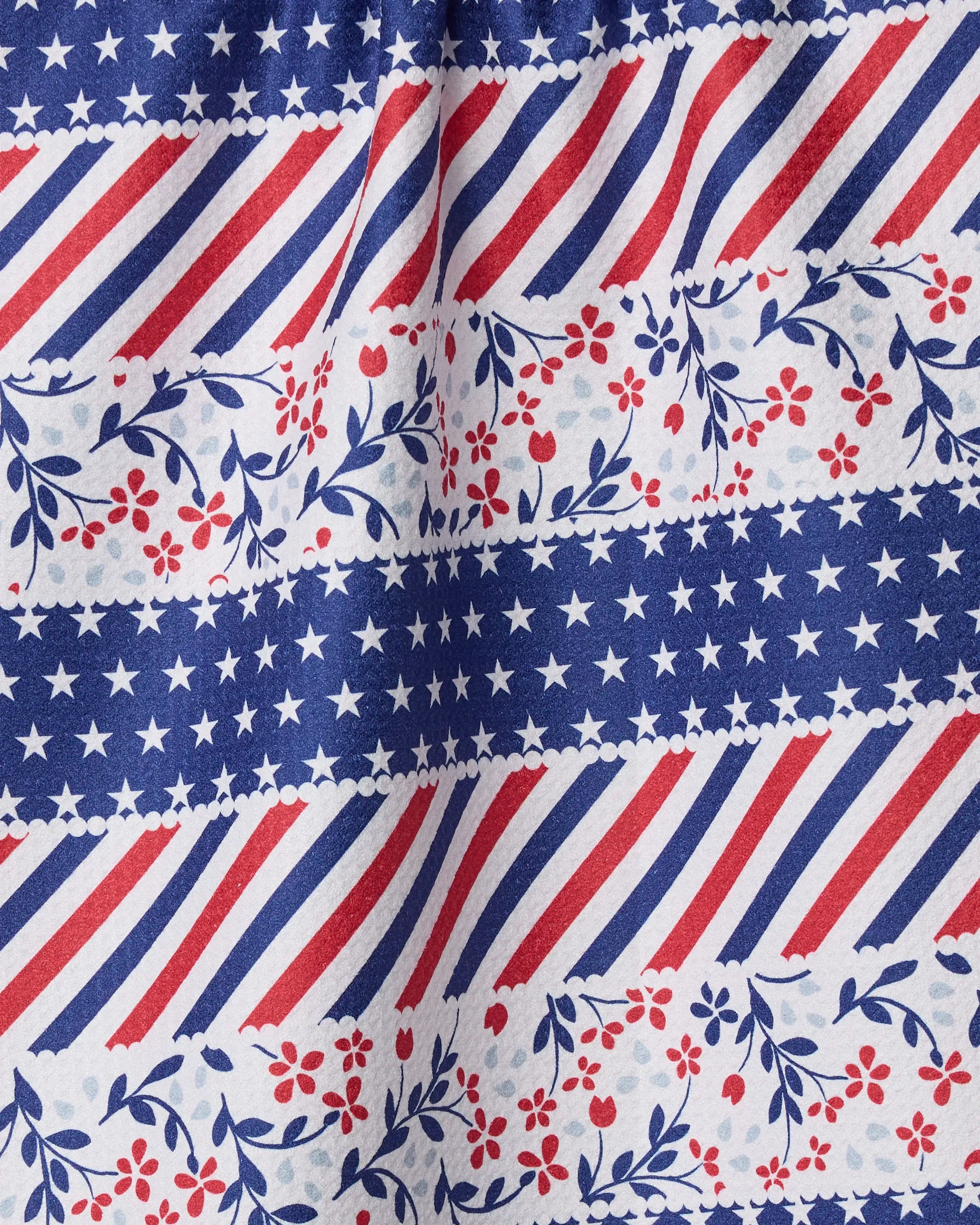 The Geometry Liberty Ribbon kitchen towel features an eco-friendly design with red, white, and blue stripes, white stars on blue, and floral accents. Soft fabric with light vertical folds makes it perfect as an absorbent dish towel.