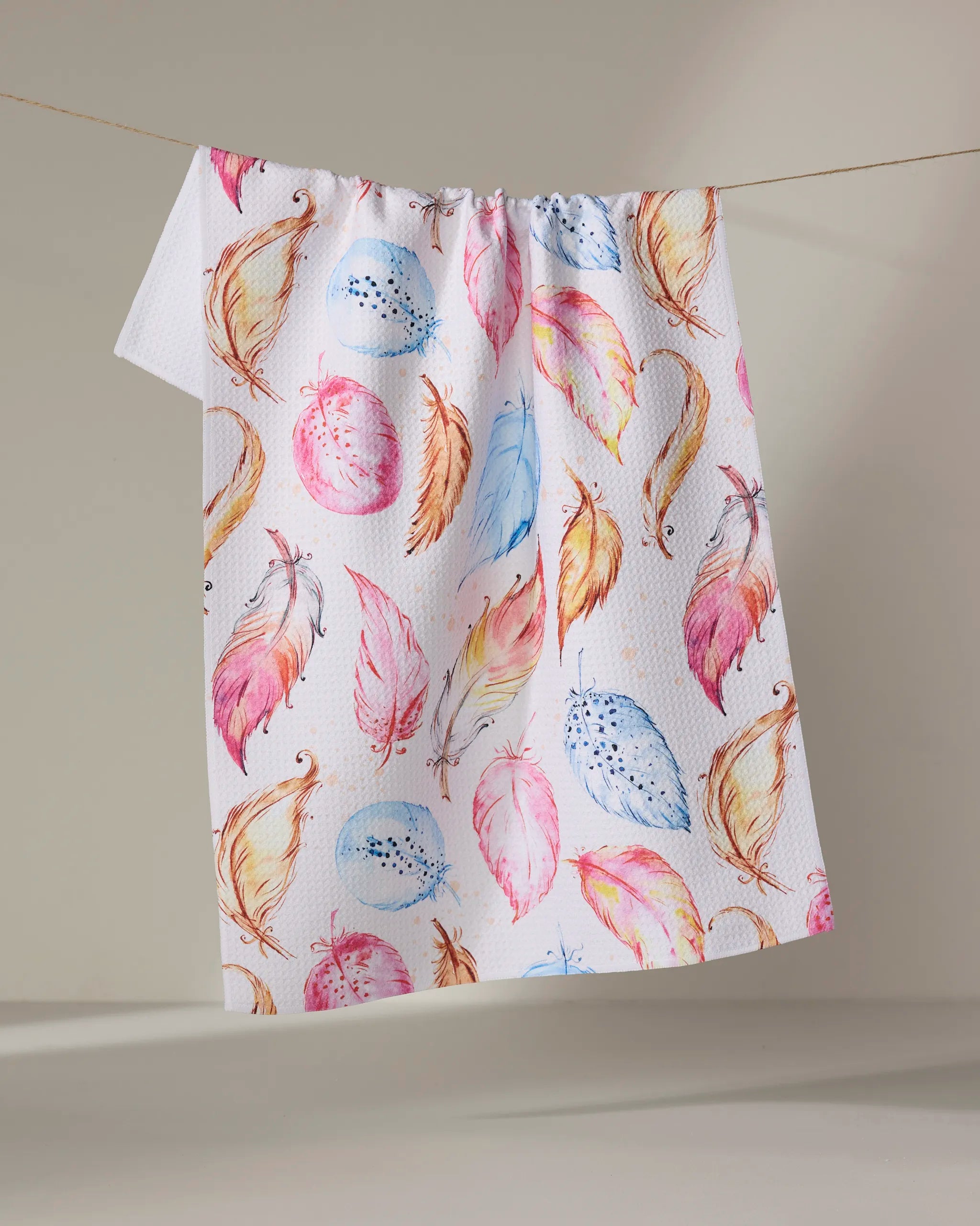 The Elena Fay "Light as a Feather" towel, ultra-absorbent and white with a vibrant feather pattern in pink, orange, yellow, and blue, is displayed hanging against a plain light background.