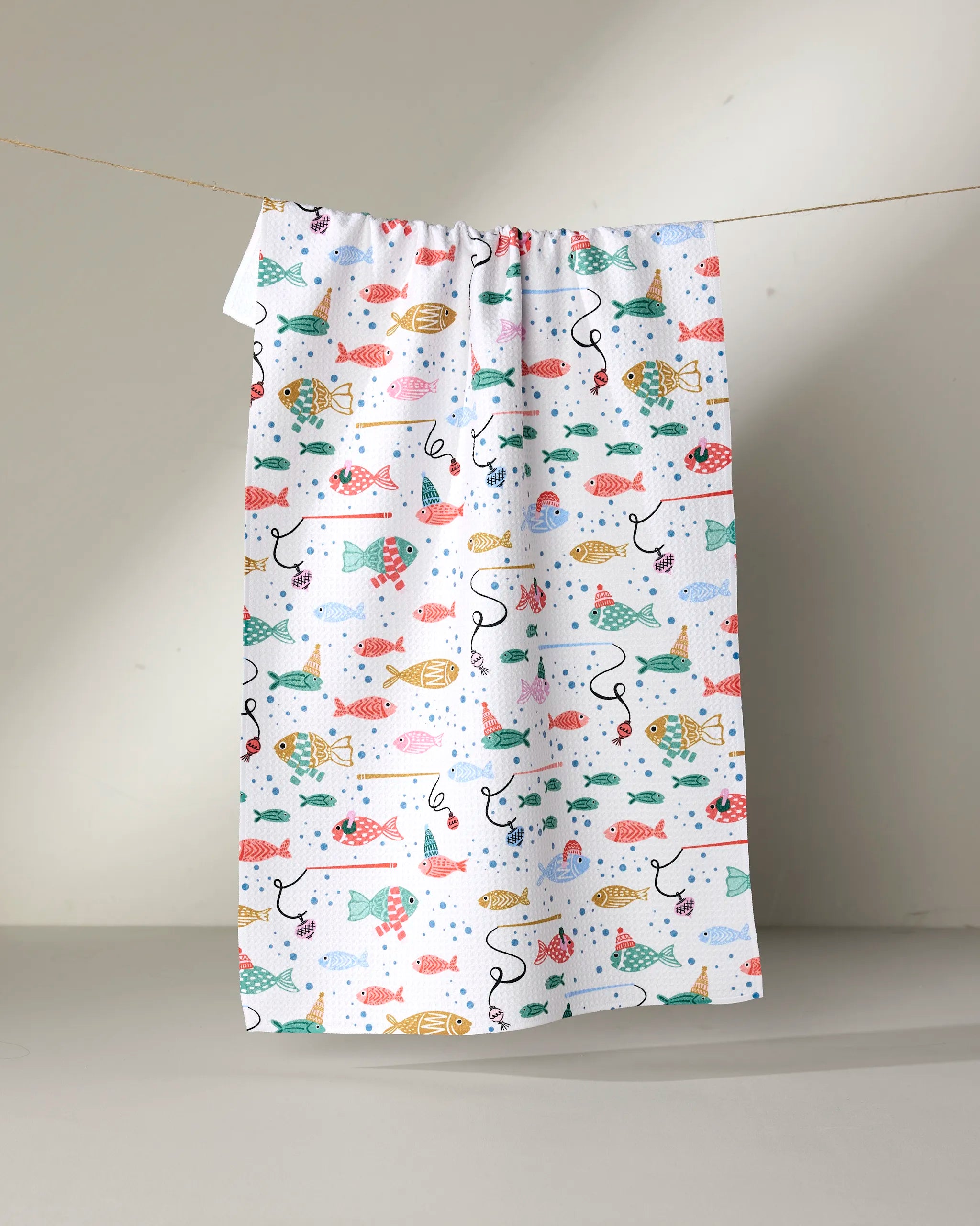 The "Light Holiday Fish" tea towel by Charlie Rowan Designs features colorful fish in red, yellow, green, and blue on white fabric, adding playful charm and a festive touch to any kitchen decor.