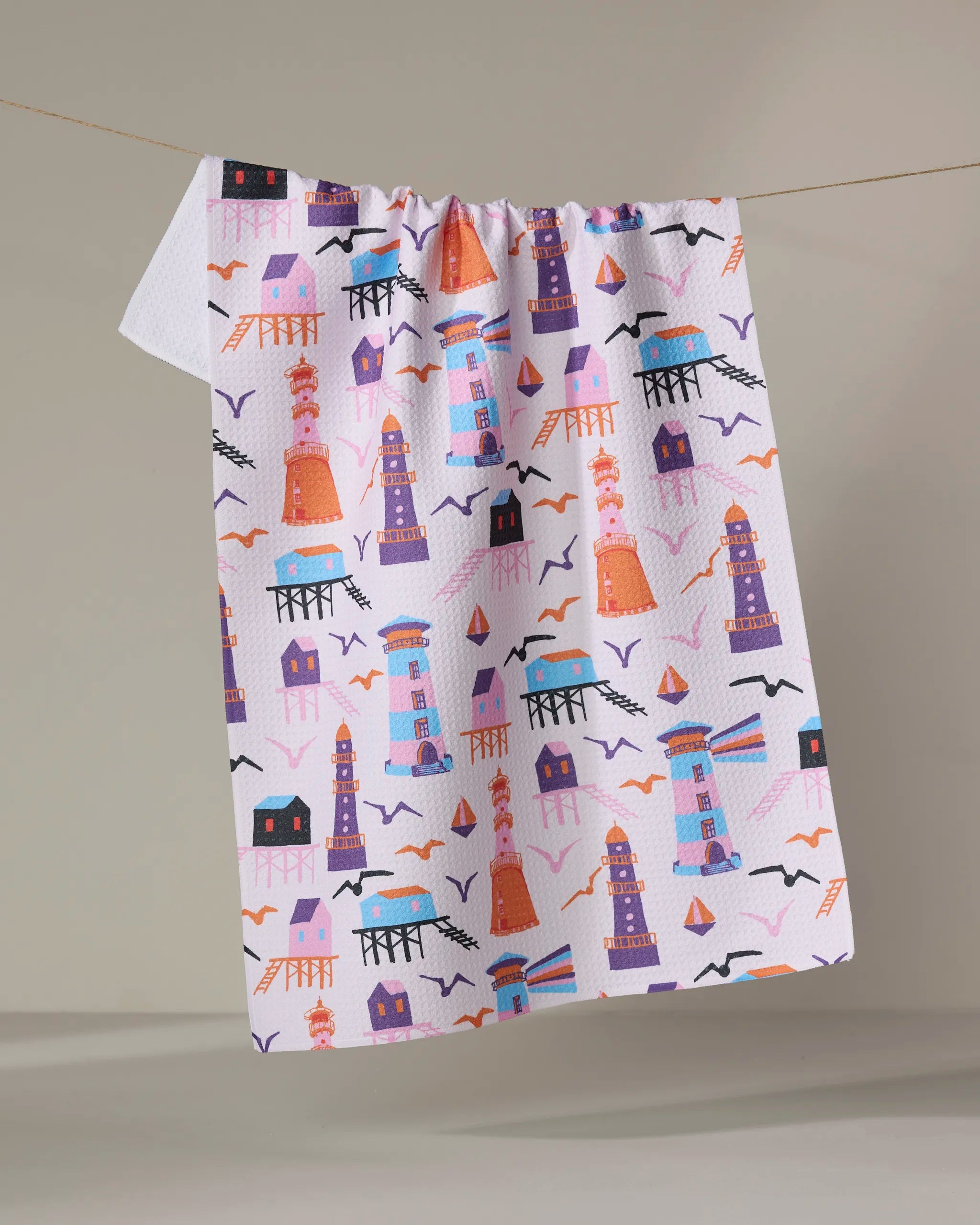 The "Lighthouse In Pink" kitchen towel by Ania Zwara Art features colorful lighthouses, seagulls, and houses in orange, purple, pink, and blue on white fabric, hanging on a clothesline against a plain background.