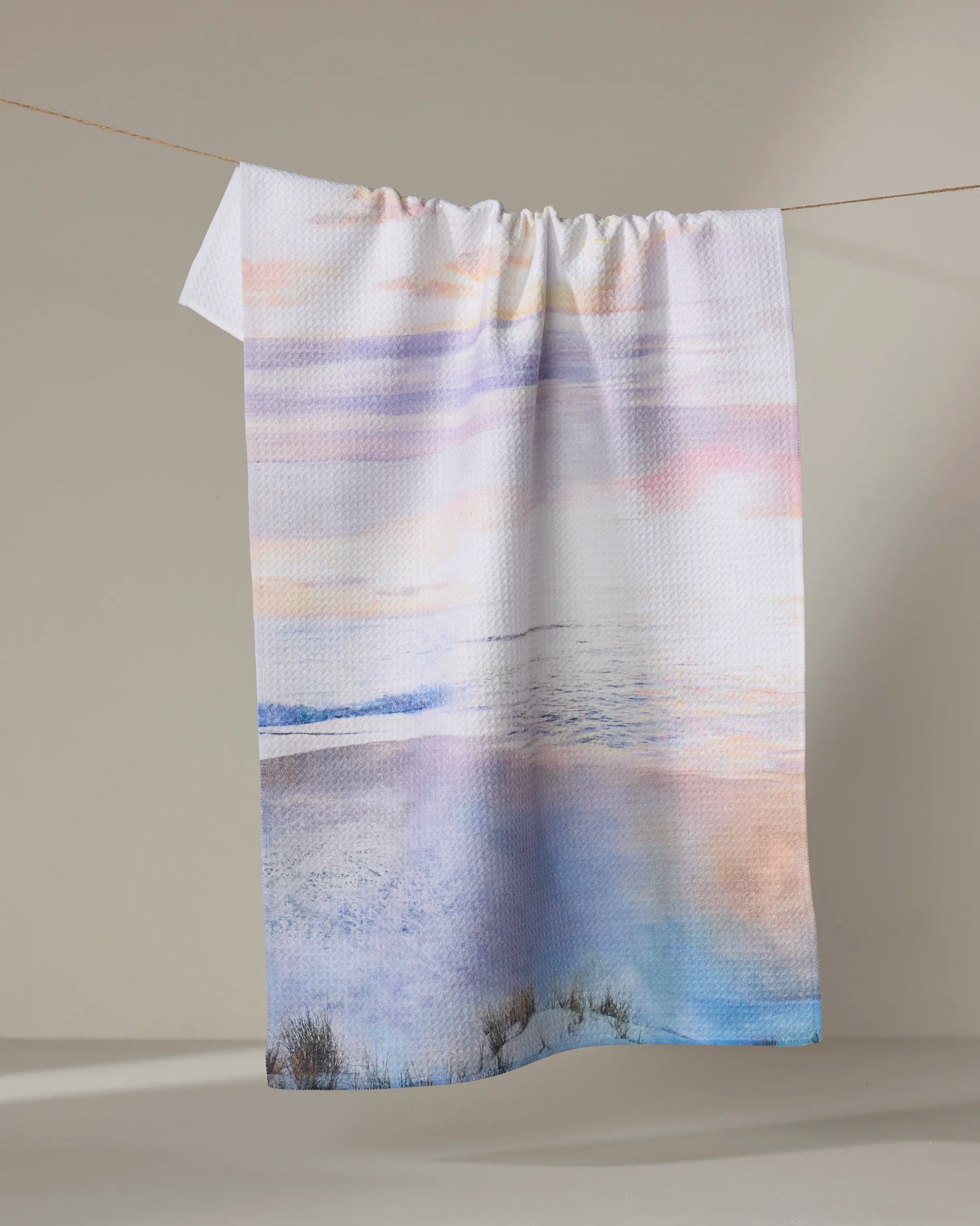 The "Lilac Coast" towel by Stefanie Bales, in pastel hues and ultra-absorbent, showcases a beach and ocean design as it hangs on a clothesline against a neutral backdrop.