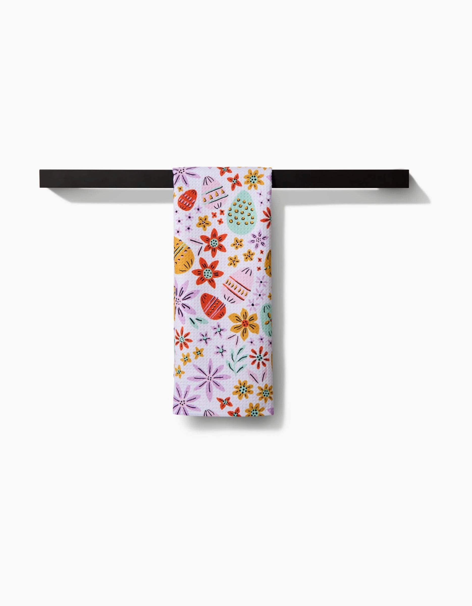 The Lilith tea towel by Amy MacCready, featuring Easter eggs and flowers, hangs neatly over a modern black towel bar against a white background.