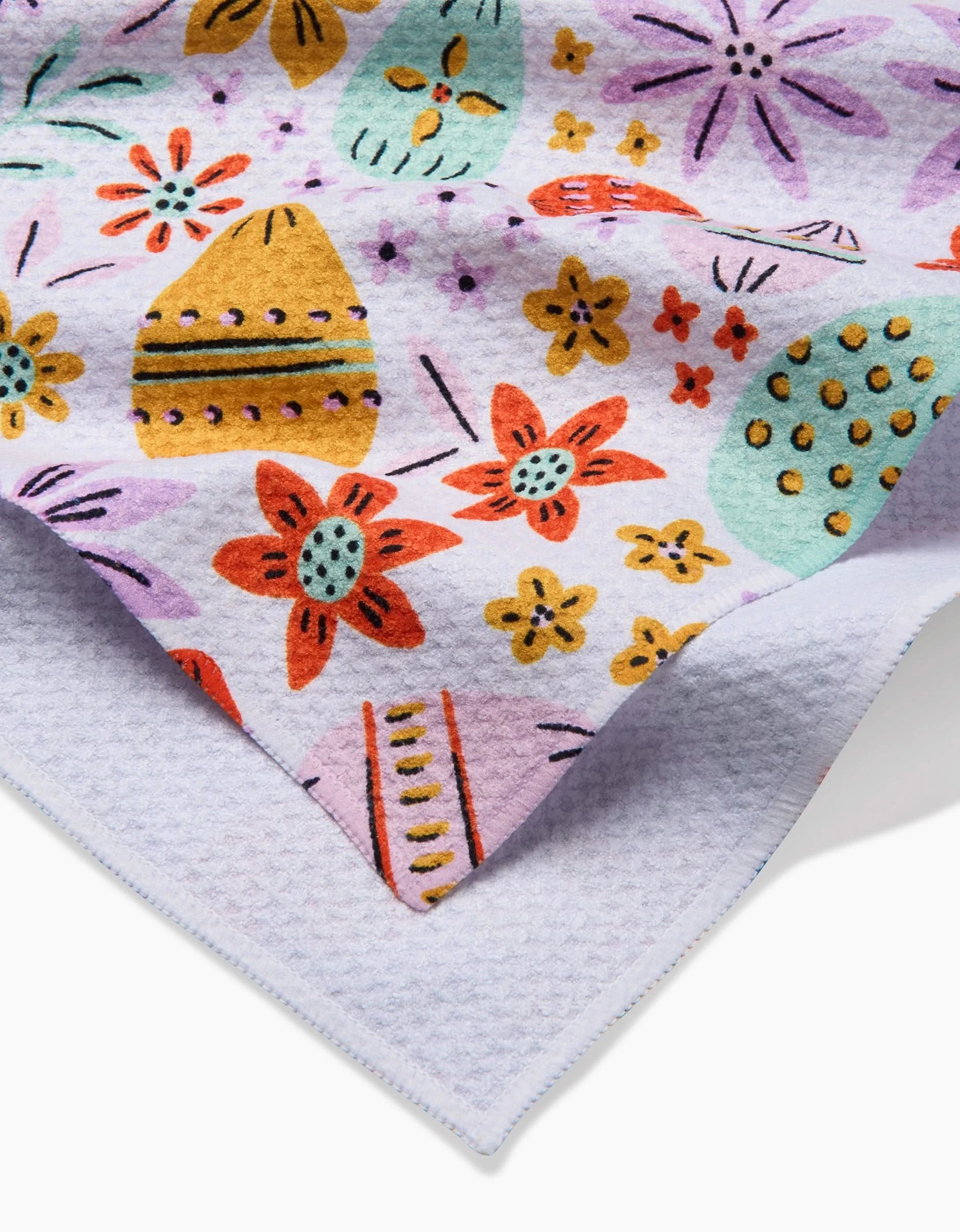 A close-up of the Lilith tea towel by Amy MacCready, featuring a vibrant floral and abstract print in red, yellow, green, and purple on white. The folded edge reveals a textured underside crafted from recycled materials.