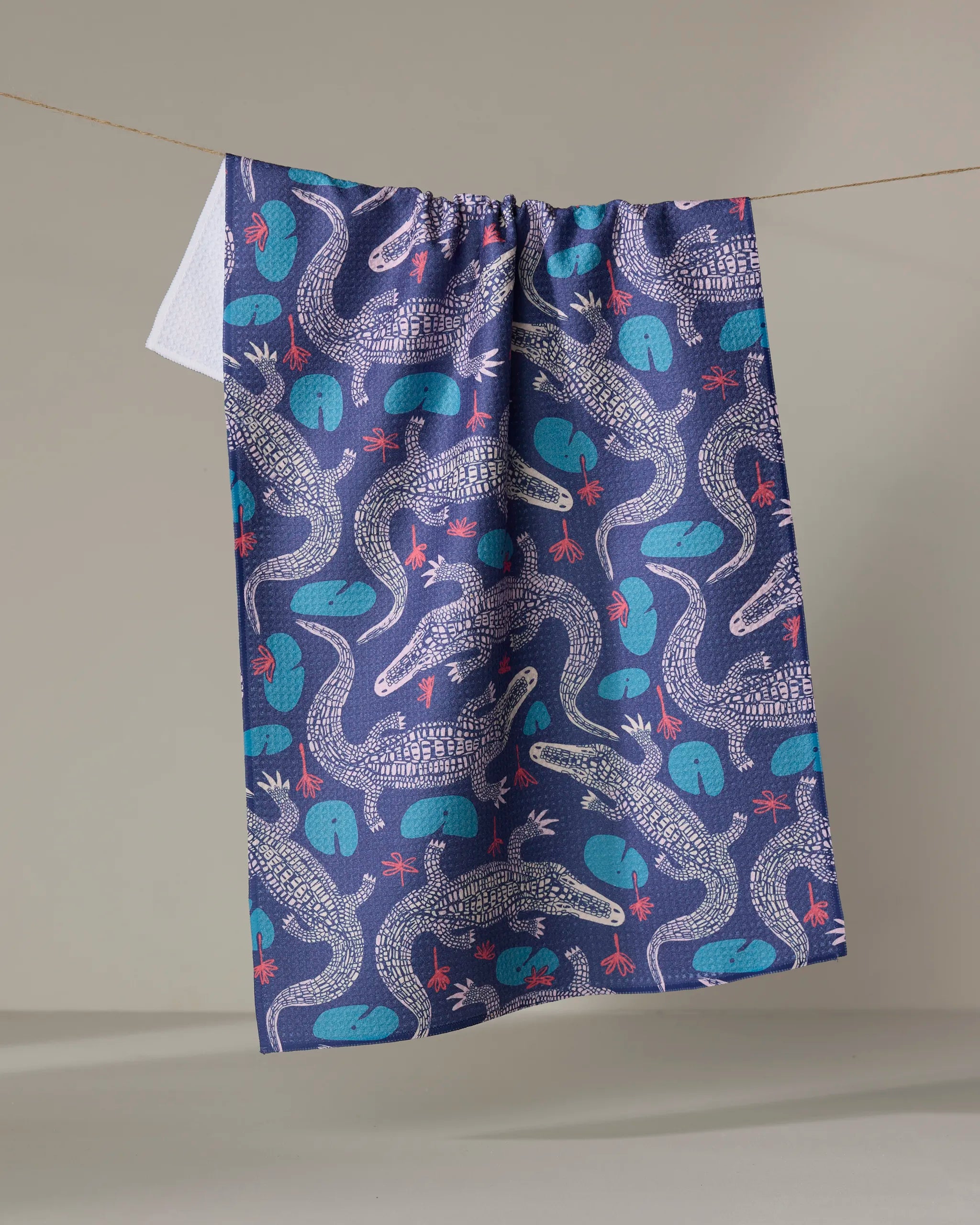 The Lillygator kitchen towel by Elisabeth Wing, made from recycled materials, features white crocodiles, teal water lilies, and red dragonflies. It hangs on a clothesline against a plain beige background.