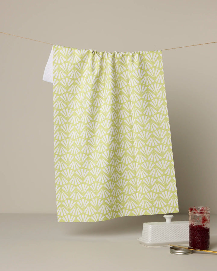 A Geometry Lime Blossom green and white patterned kitchen towel hangs on a line above a butter dish, an open jar of red jam, and its lid, all set on a light surface with a neutral background.