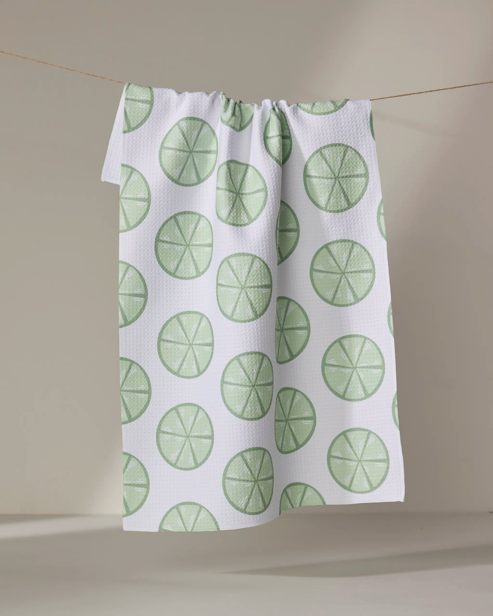 The “Lime Doodles” tea towel by Letters By Shells, featuring green lime slice patterns on white, hangs over a clothesline with a neutral backdrop—an absorbent and eco-friendly kitchen essential.