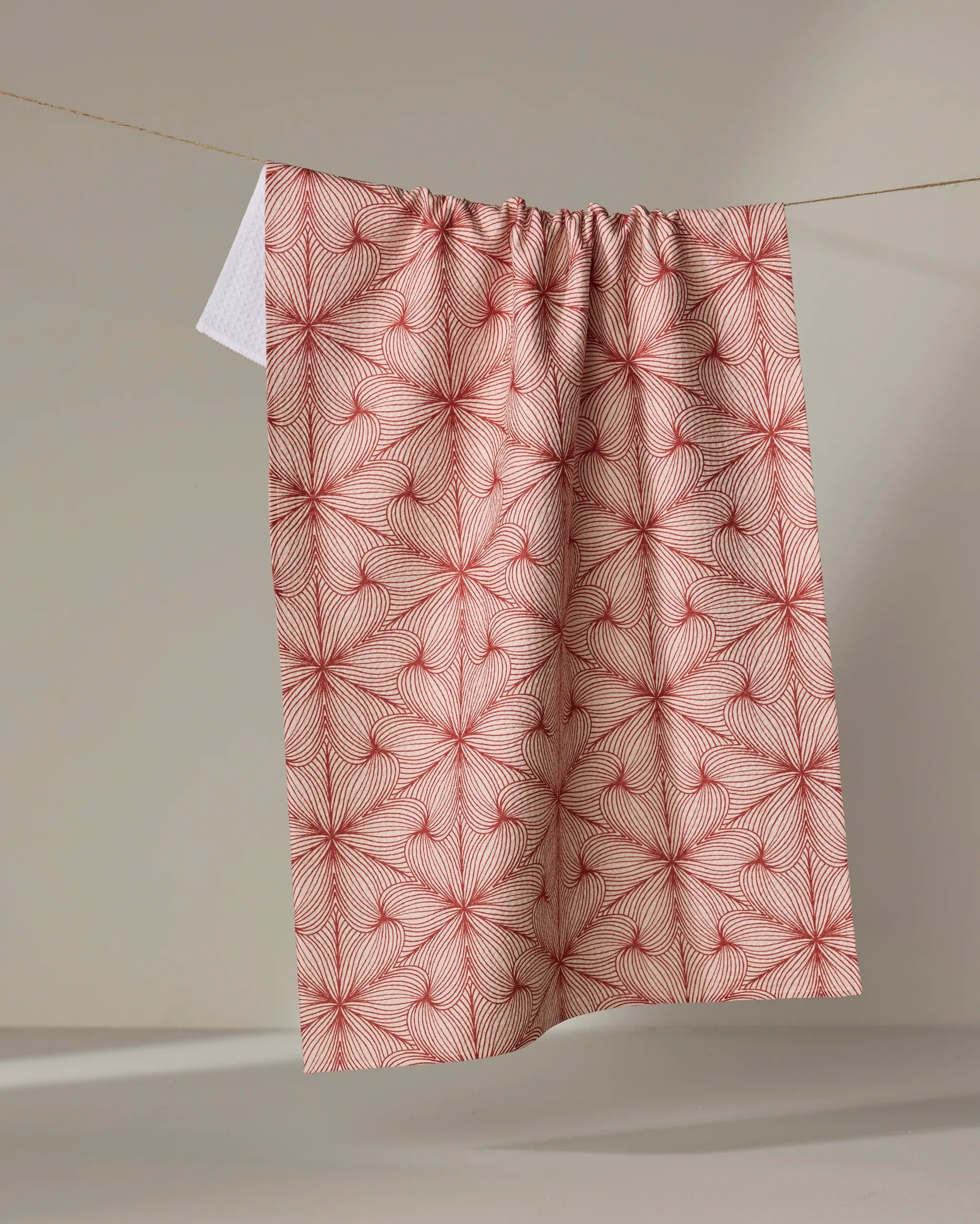 The "Lined" tea towel by Marta Harvey, featuring a red geometric, leaf-inspired pattern, is draped over a clothesline against a neutral background.