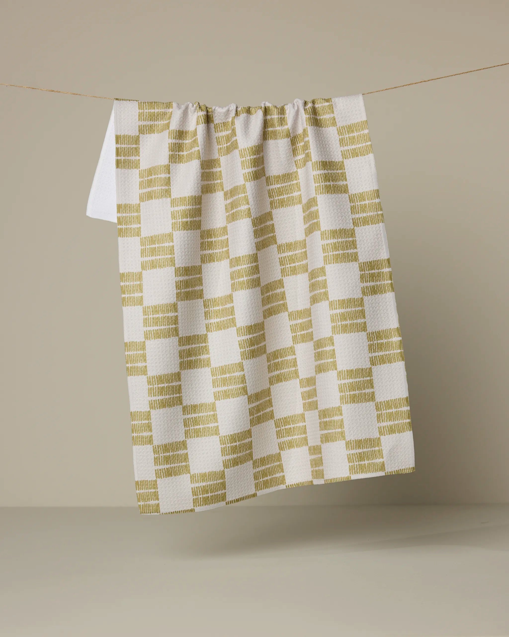 The Linnea tea towel by Anna Christian, made from ultra-absorbent recycled materials in a yellow checkered pattern, hangs from a thin string against a neutral beige background.