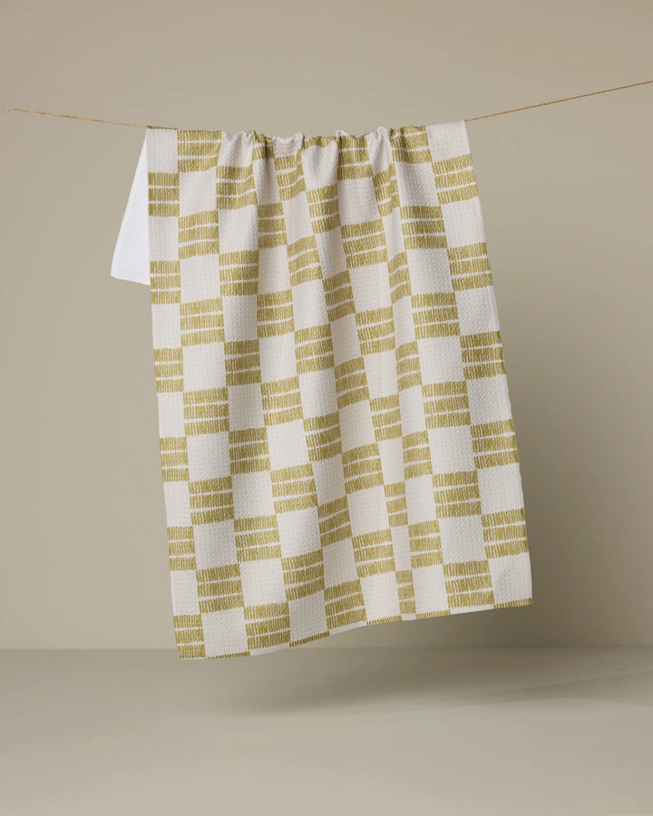 The Linnea tea towel by Anna Christian, made from ultra-absorbent recycled materials in a yellow checkered pattern, hangs from a thin string against a neutral beige background.