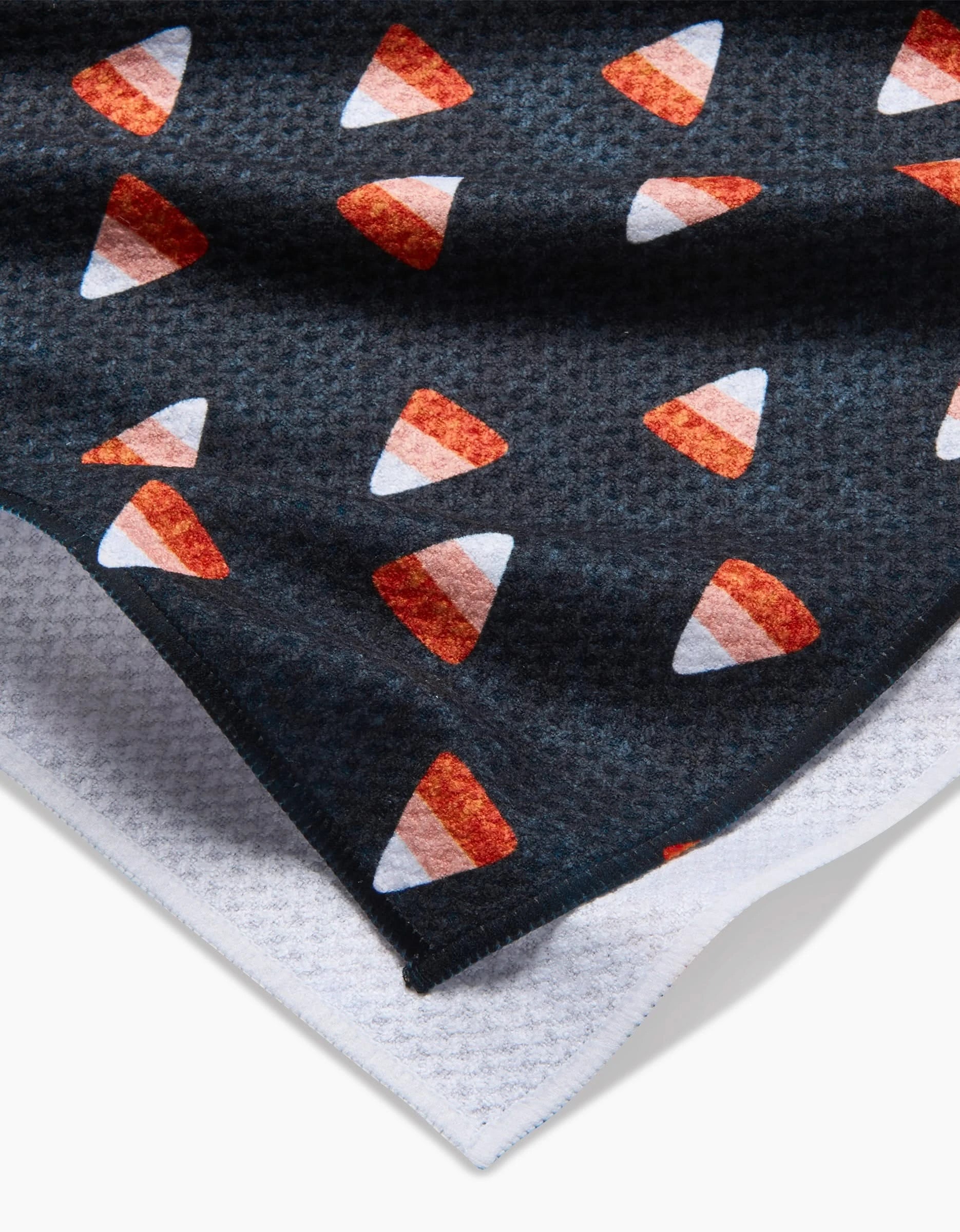 The Linnet kitchen tea towel by Amy MacCready features a dark, textured fabric with a repeating candy corn pattern in white, orange, and yellow. A folded corner reveals the white underside made from recycled materials.