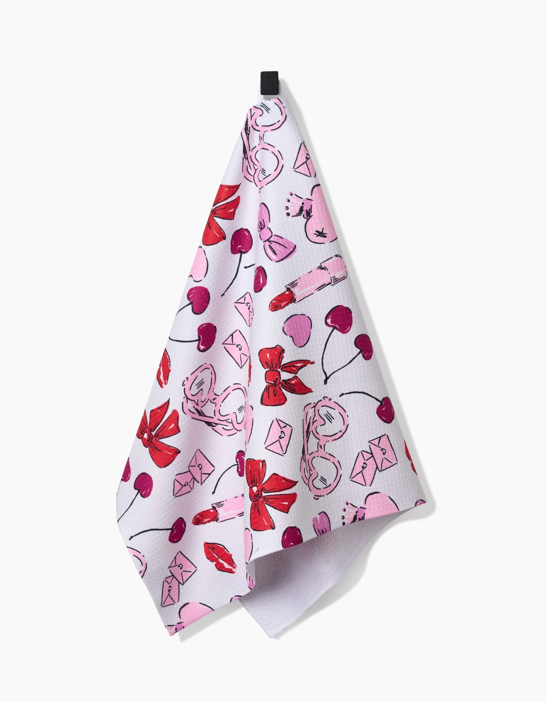 The Lip Smacking Good kitchen towel by Hufton Studio, featuring playful pink and red cherries, bows, flowers, and envelopes on white recycled fabric, hangs from a black hook against a plain white background.