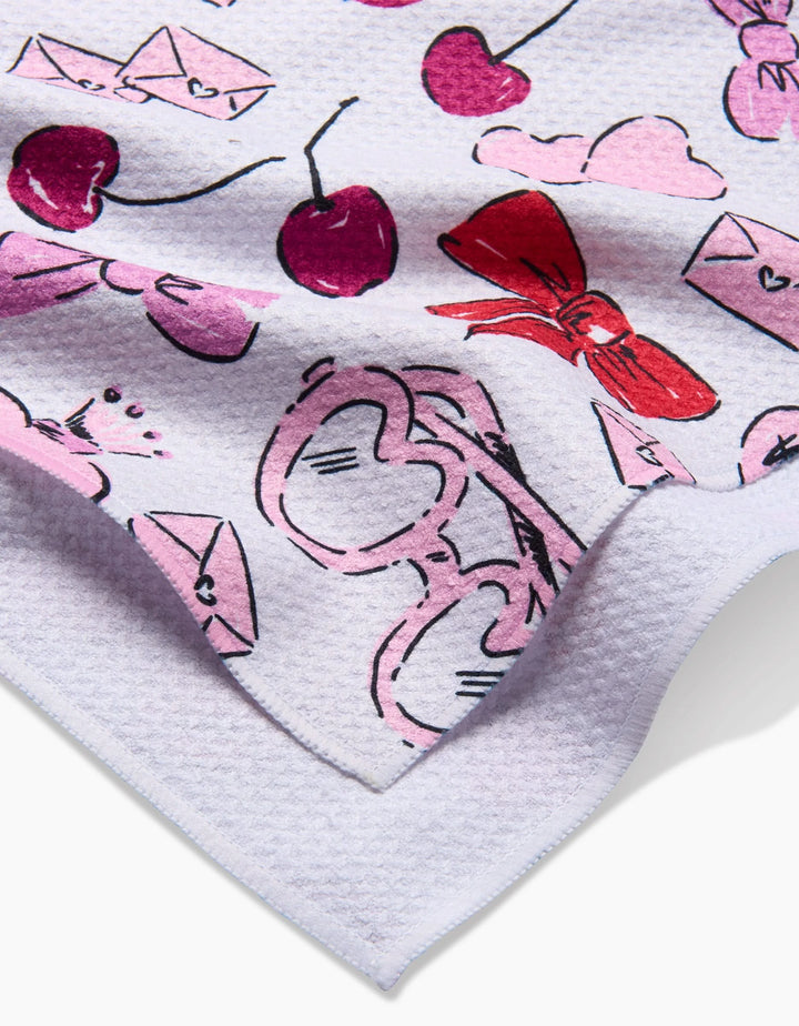 A close-up of Lip Smacking Good, a white textured kitchen towel by Hufton Studio, with playful pink and red hearts, cherries, bows, sunglasses, clouds, and envelopes. The recycled tea towel is slightly folded at one corner.