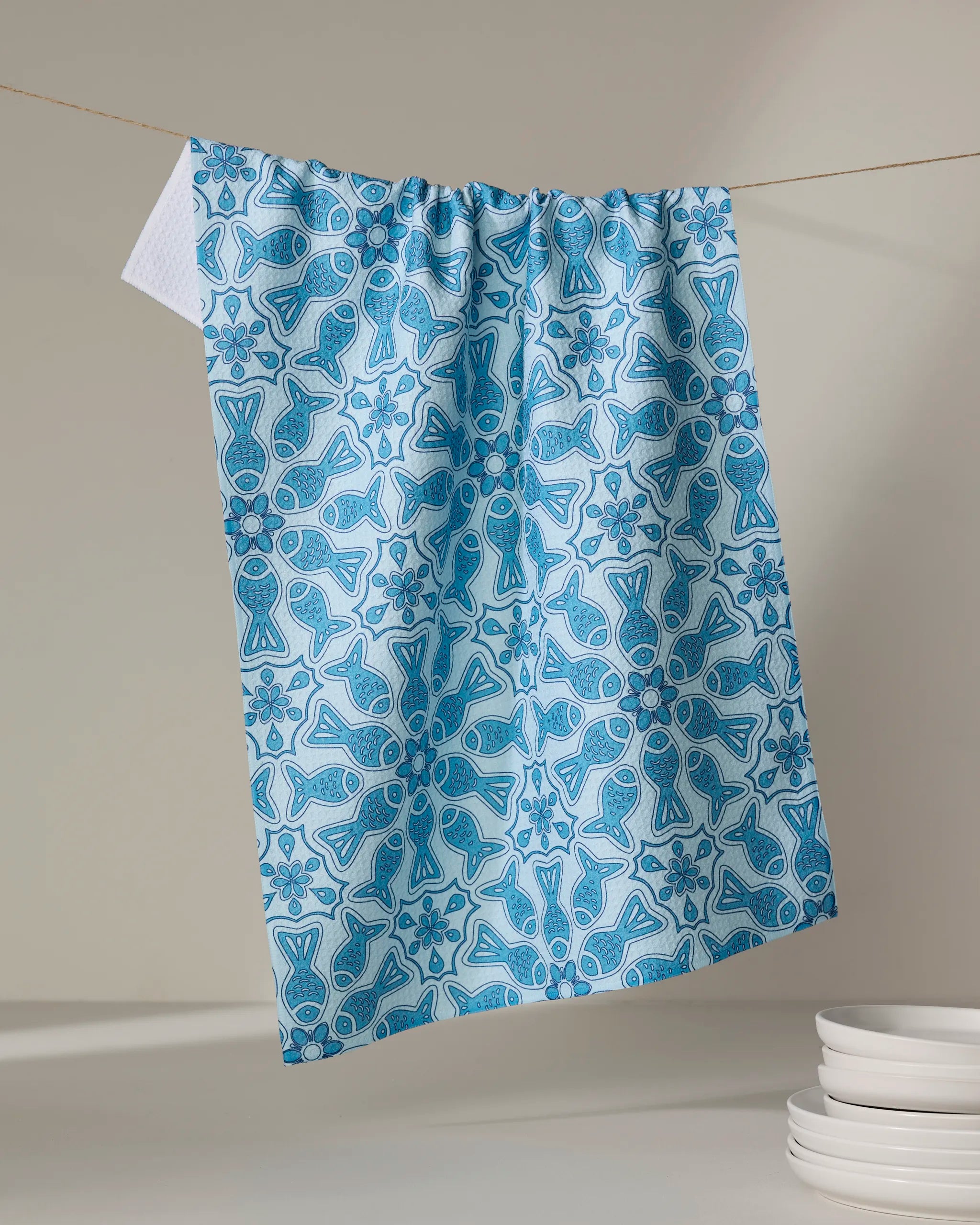 The Little Fish Mandala kitchen tea towel by Marta Harvey, made from recycled materials, features a blue and white fish and floral pattern. Shown on a clothesline beside white plates against a neutral background.