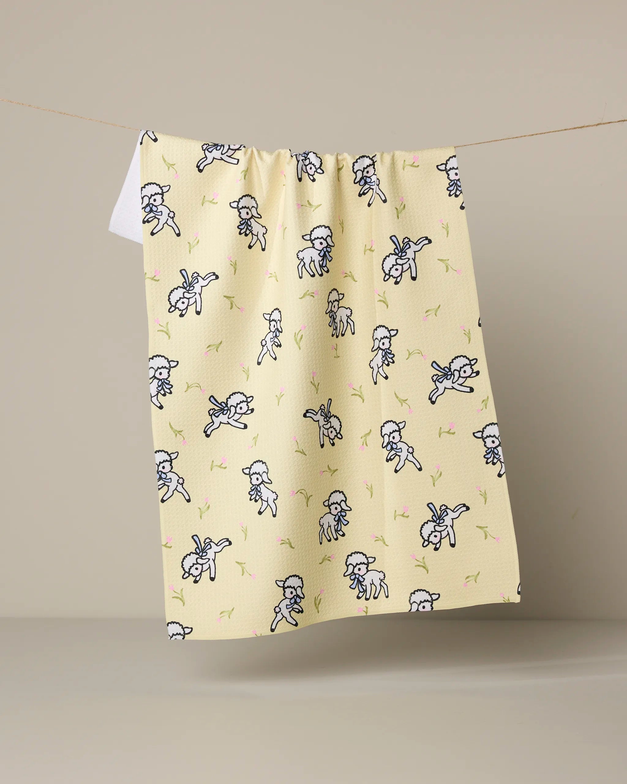 The "Little Lamb" kitchen towel by Geometry, featuring cartoon angels with trumpets and pink and green accents, is draped over a line against a plain beige background.