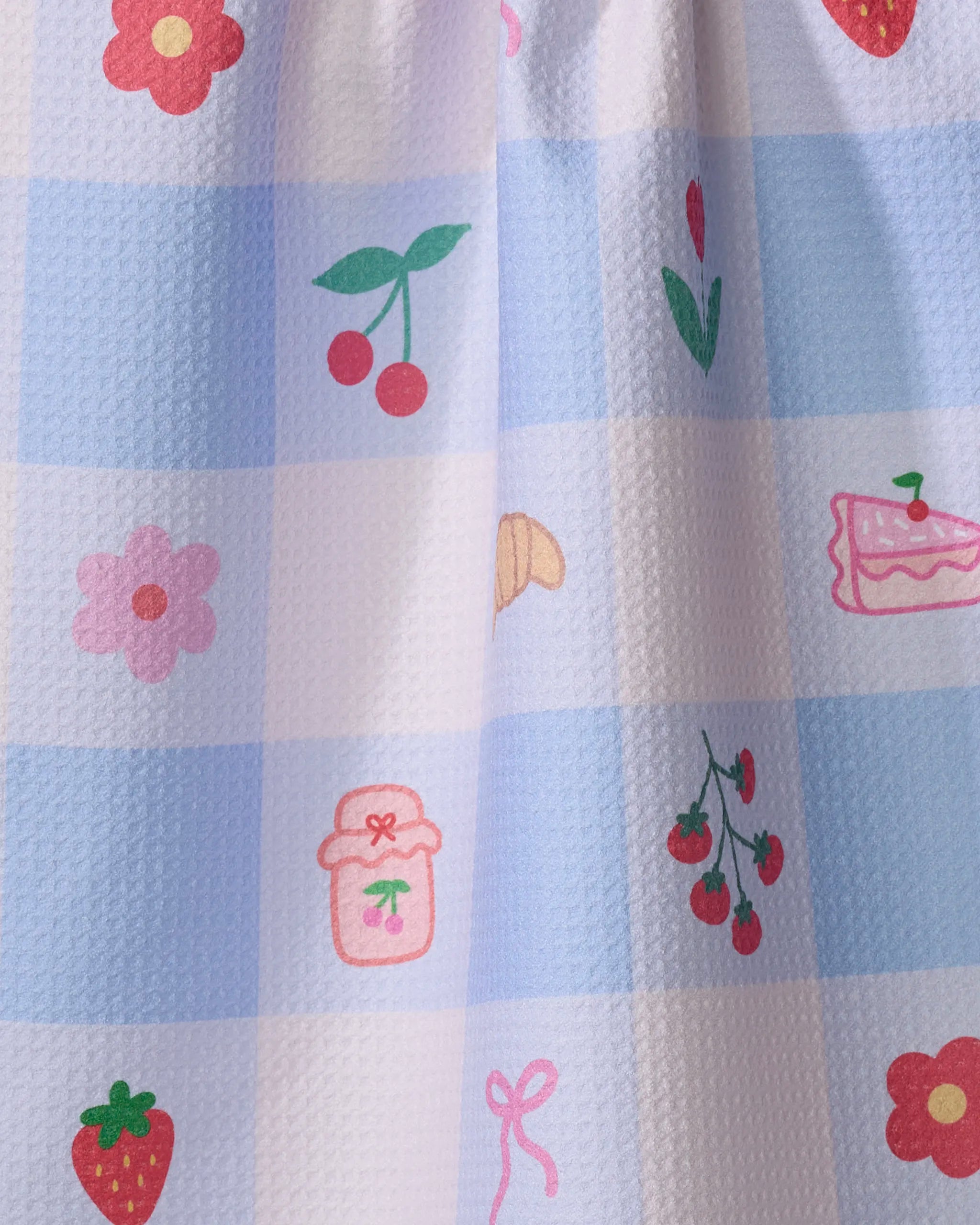 The Little Picnic Things tea towel by Letters By Shells features blue and white checks with cartoon cherries, strawberries, flowers, a cake slice, jar, pink bow, and leaves. Made from recycled materials for an eco-friendly touch.