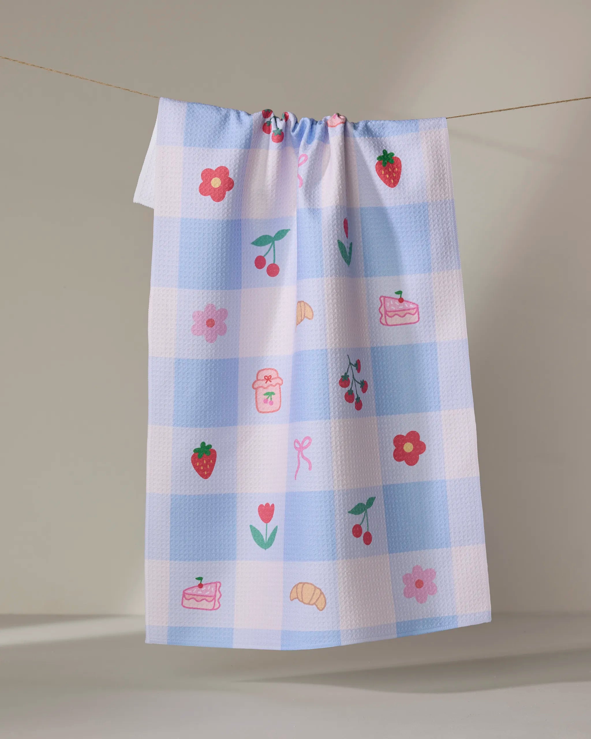 The "Little Picnic Things" tea towel by Letters By Shells features blue and white checks with charming illustrations of flowers, strawberries, cherries, a croissant, jam jars, cake slices, and bows.