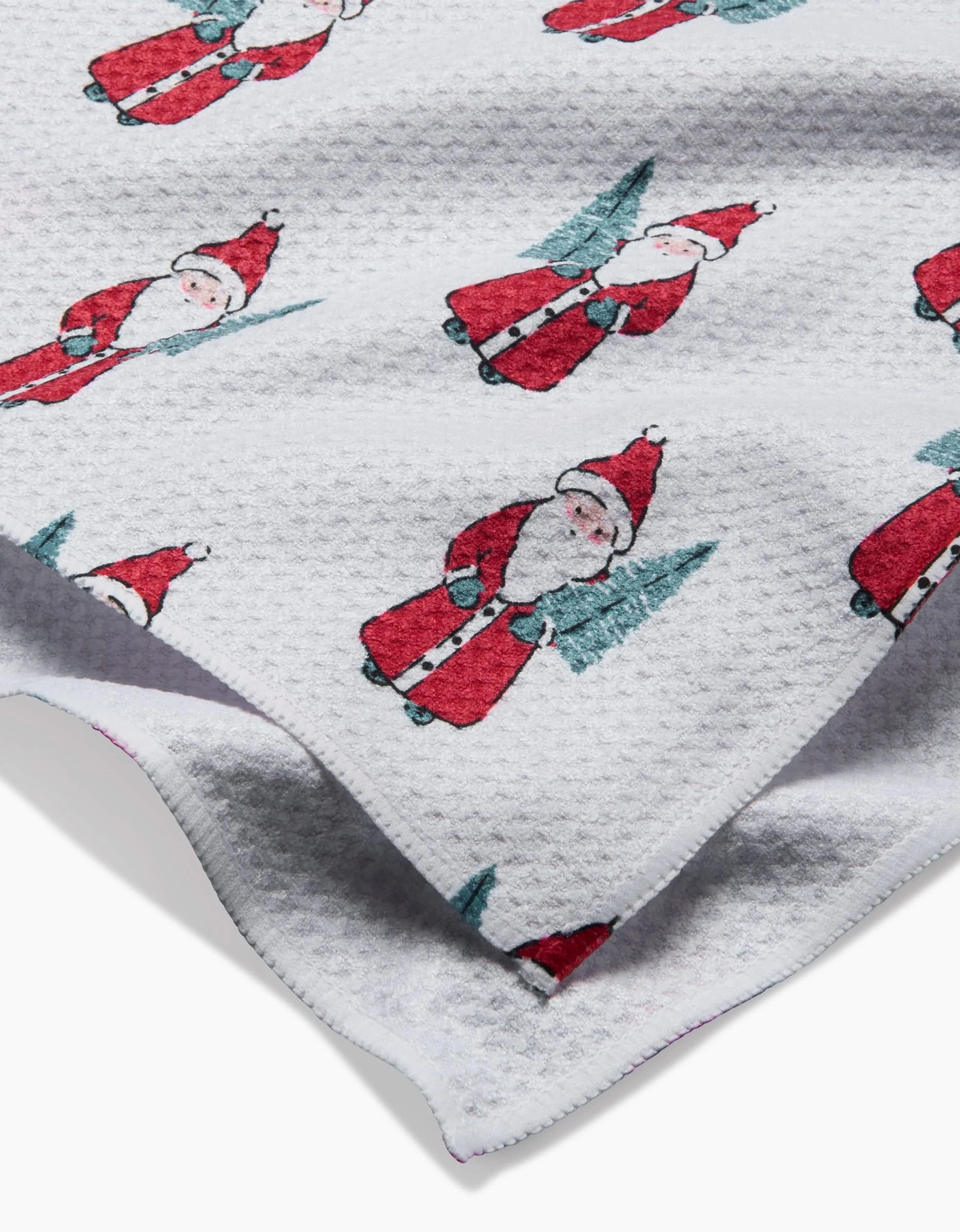The Little Santa recycled kitchen towel by Hufton Studio features a textured white design with a repeating Santa Claus holding a green Christmas tree, in red hats and coats, shown draped and partially folded.