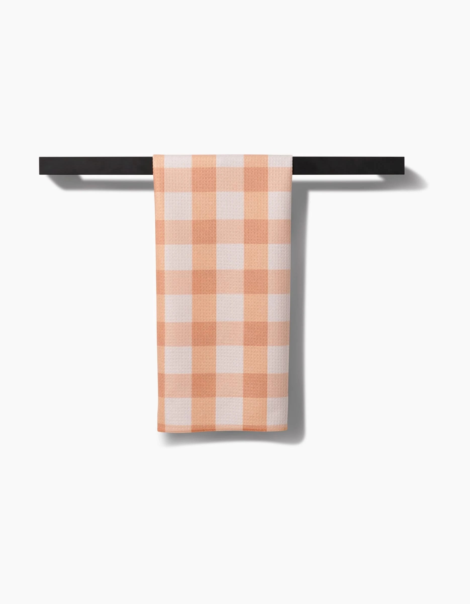 The Lorraine tea towel by Kelsi Fullmer, featuring a peach and white gingham check pattern and made from recycled materials, hangs neatly over a black bar with a white background.