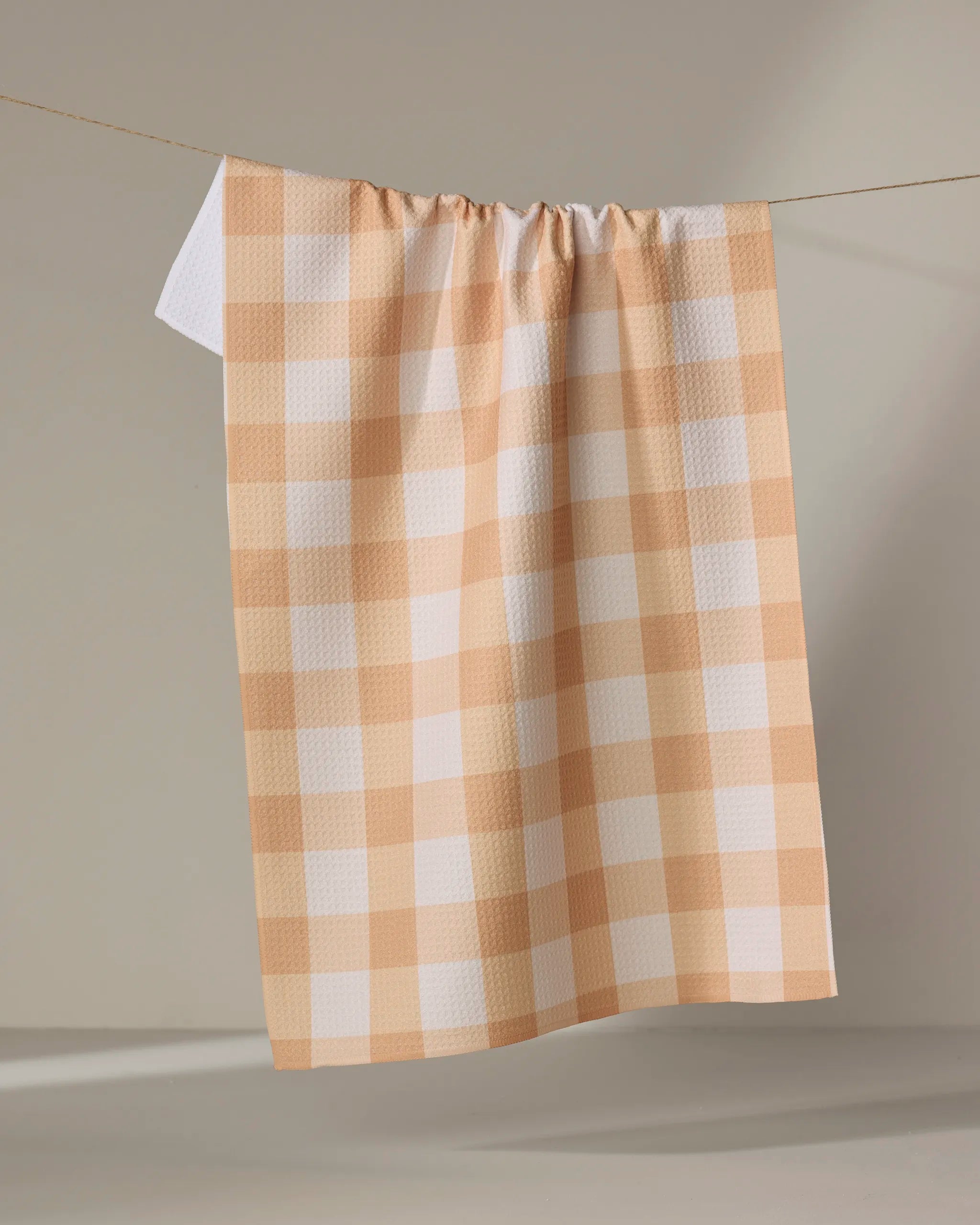 The Lorraine kitchen tea towel by Kelsi Fullmer, featuring a beige and white checkered design and made from recycled materials, hangs from a string against a plain light background.
