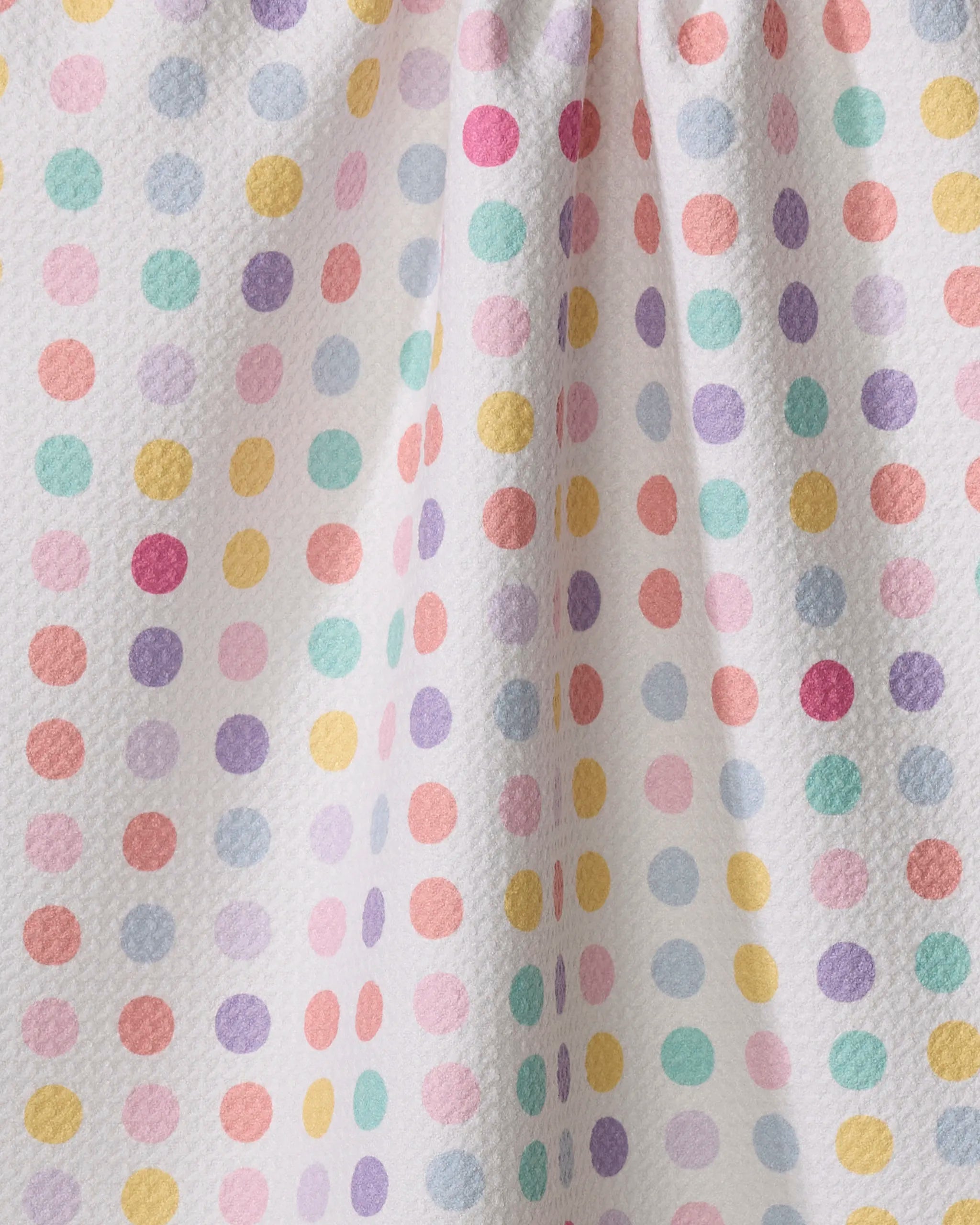 The "Lots of Dots" tea towel by Rebecca Bobko features a white textured fabric with pastel polka dots in pink, yellow, orange, green, blue, and purple. Slight gathering adds gentle folds and subtle shadows.