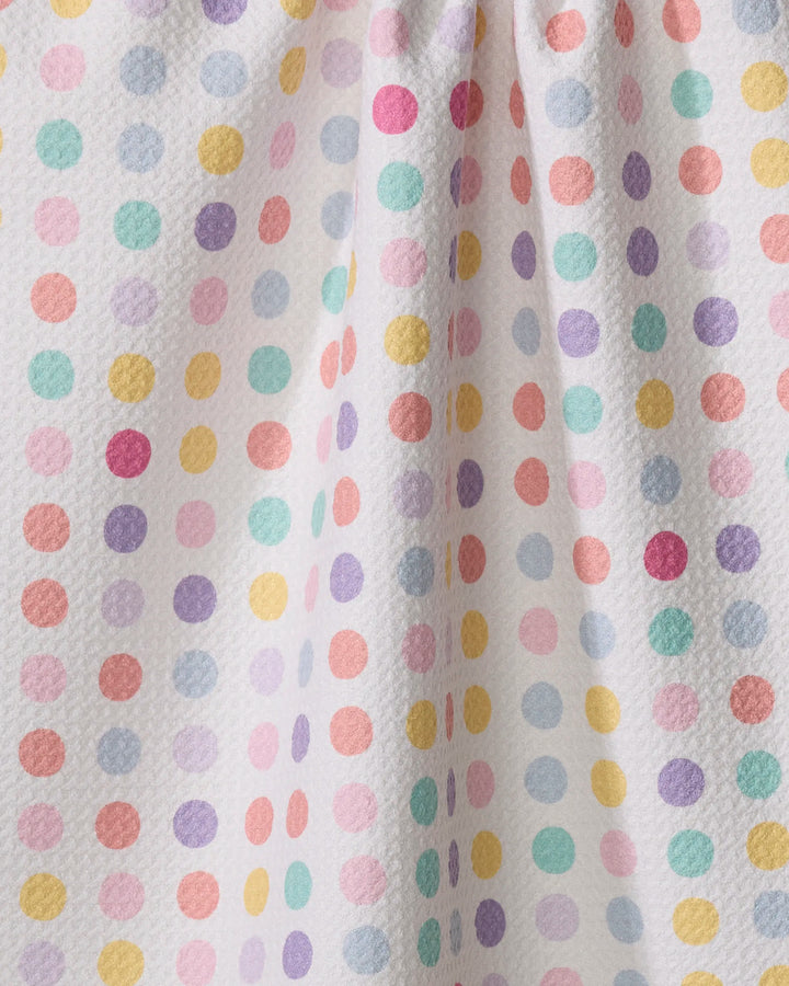 The "Lots of Dots" tea towel by Rebecca Bobko features a white textured fabric with pastel polka dots in pink, yellow, orange, green, blue, and purple. Slight gathering adds gentle folds and subtle shadows.