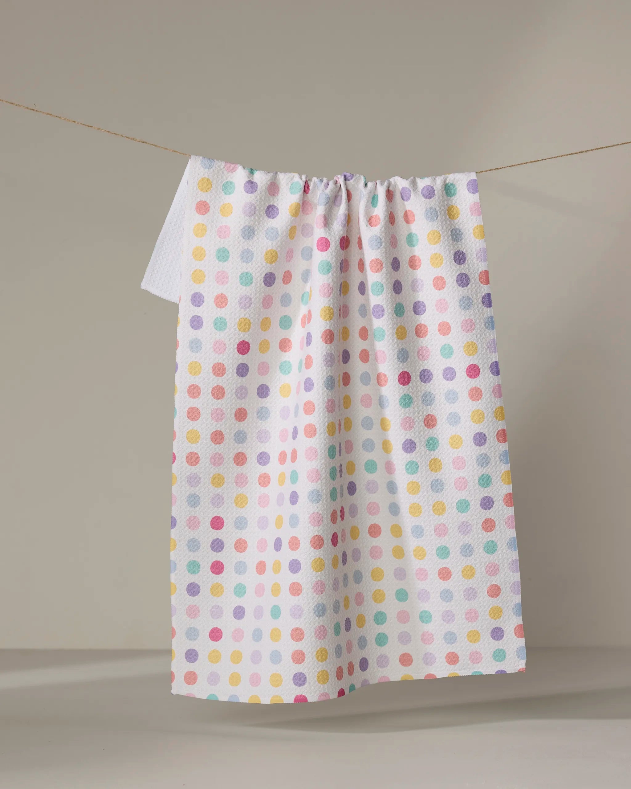 The "Lots of Dots" dish towel by Rebecca Bobko, featuring a pastel polka dot pattern on white fabric, hangs on a clothesline against a neutral background.