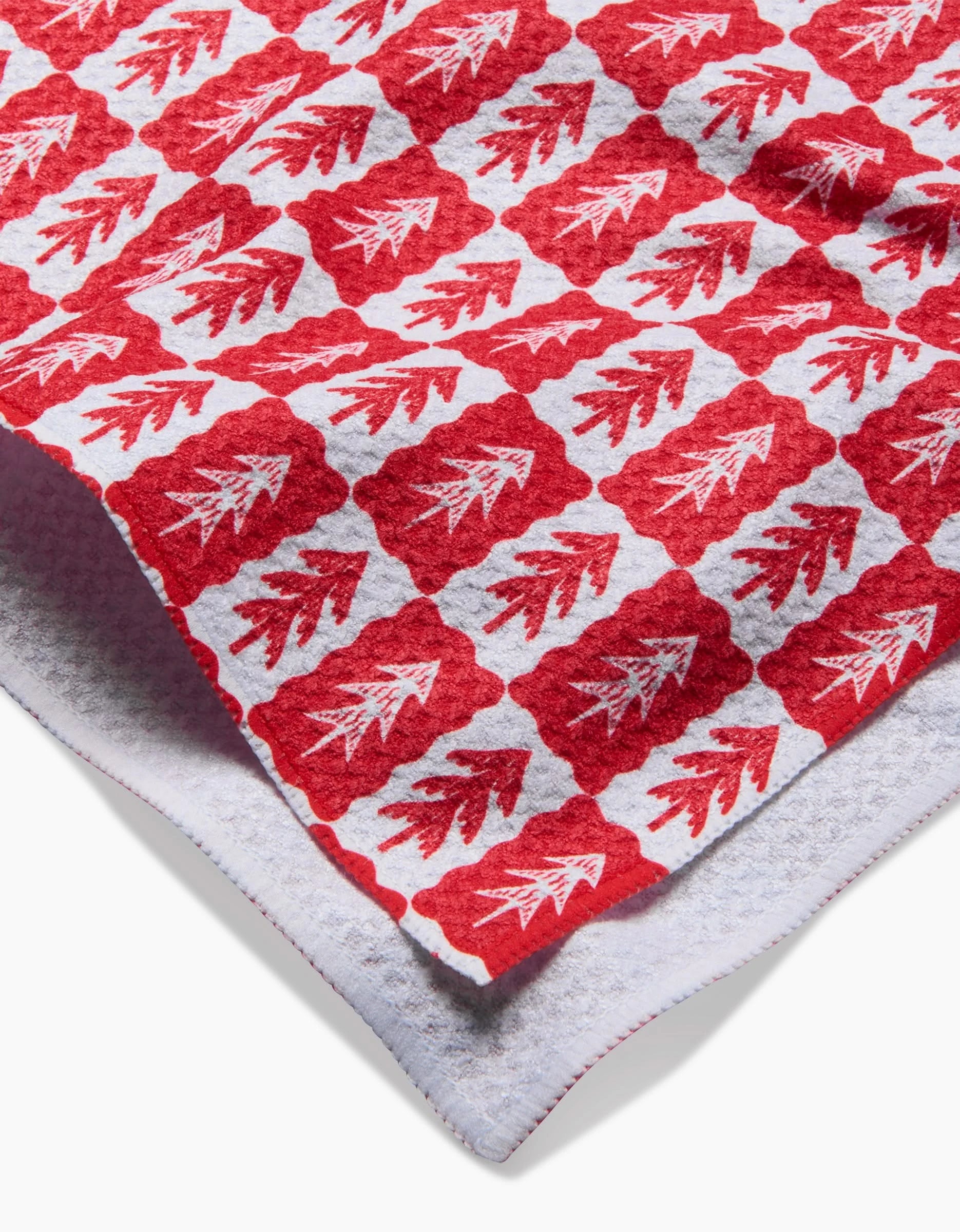 A close-up of the Roxy Marj Louis Pines tea towel shows its red and white pine tree pattern on a textured surface, with one corner folded to reveal the reverse side.