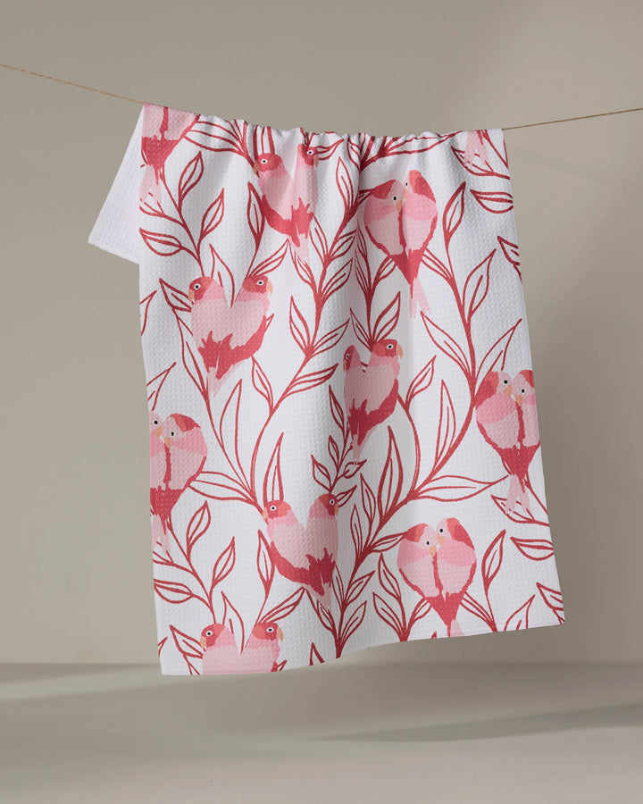 The Love Birds kitchen towel by Hufton Studio, featuring a pink bird-on-branches pattern on white recycled fabric, hangs draped over a clothesline against a plain beige background.