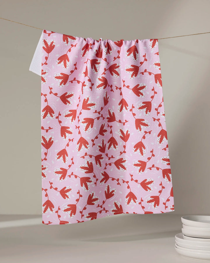 The Love Blooms tea towel by Charlie Rowan Designs, with its red and white floral pattern on pink, hangs on a clothesline brightening the space, while stacked white dishes rest nearby.