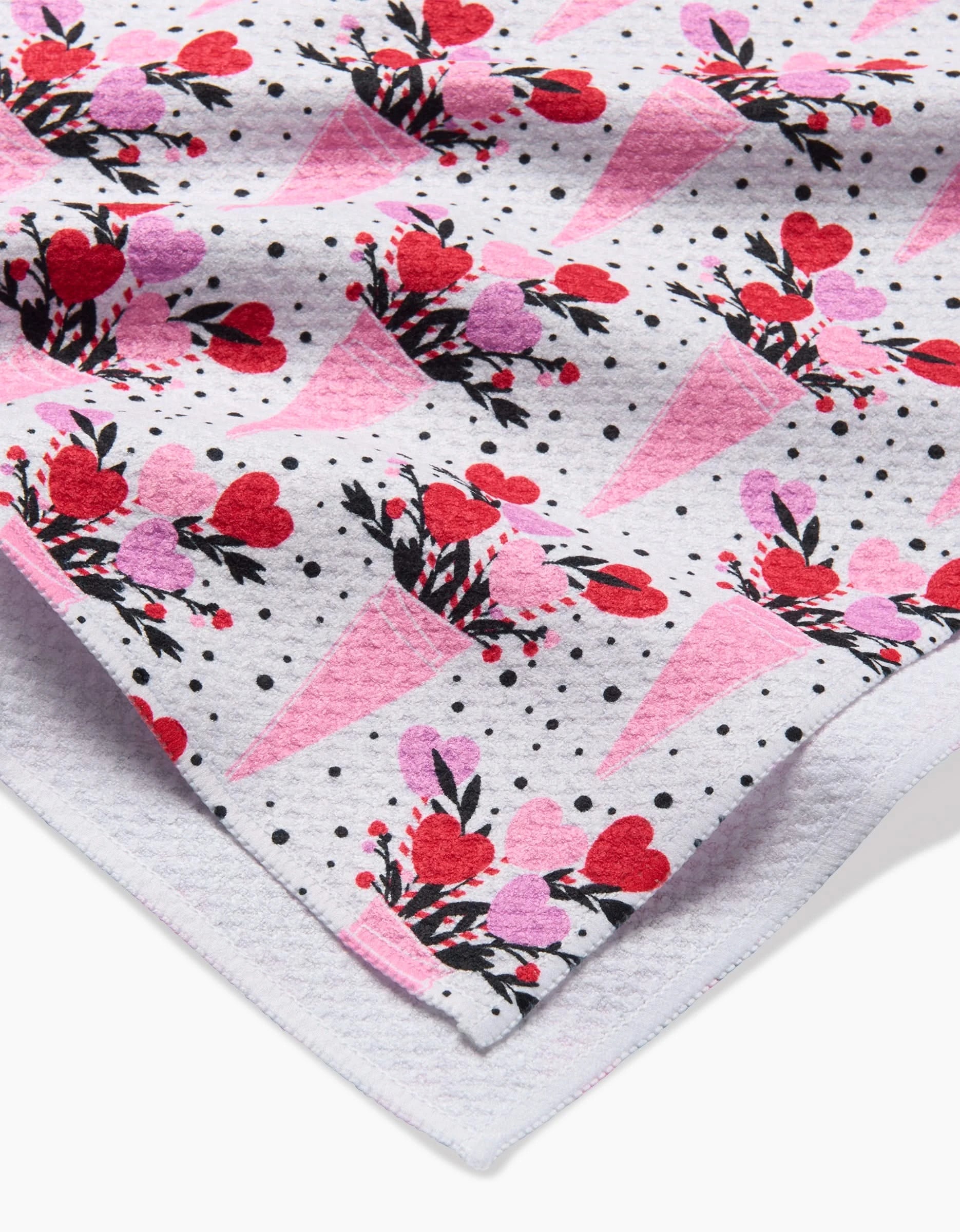 A close-up of the Love Bouquet recycled kitchen towel by Charlie Rowan Designs, in soft light gray with a pattern of pink cones holding red and pink heart flowers, black leaves, and black polka dots.