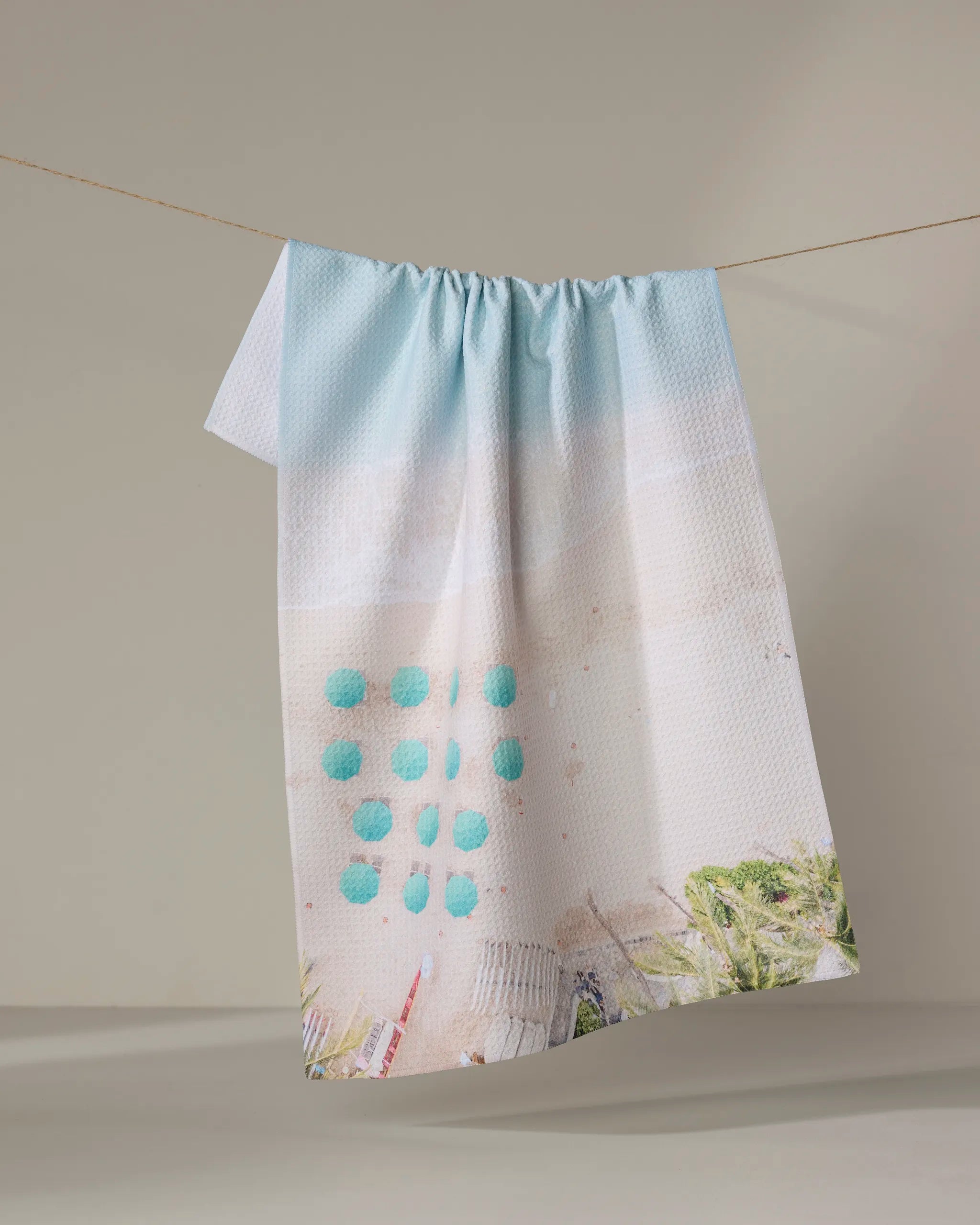 The "Love From Hawaii" dish towel by Kait Hanson, featuring a light blue and white design with teal dots and greenery accents, hangs on a clothesline against a neutral background.