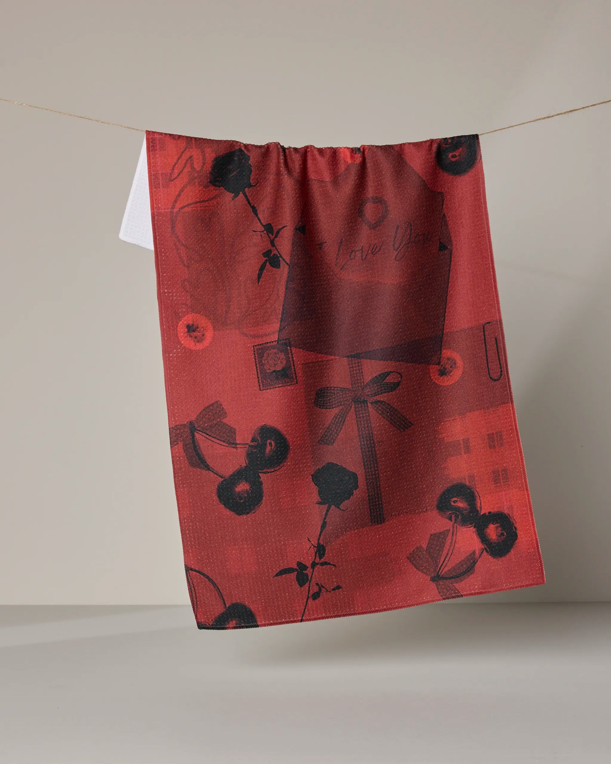 The Geometry Love Journal tea towel, made from recycled materials, displays black and dark red roses, cherries, hearts, and handwritten "I love you" designs as it hangs on a clothesline against a neutral background.