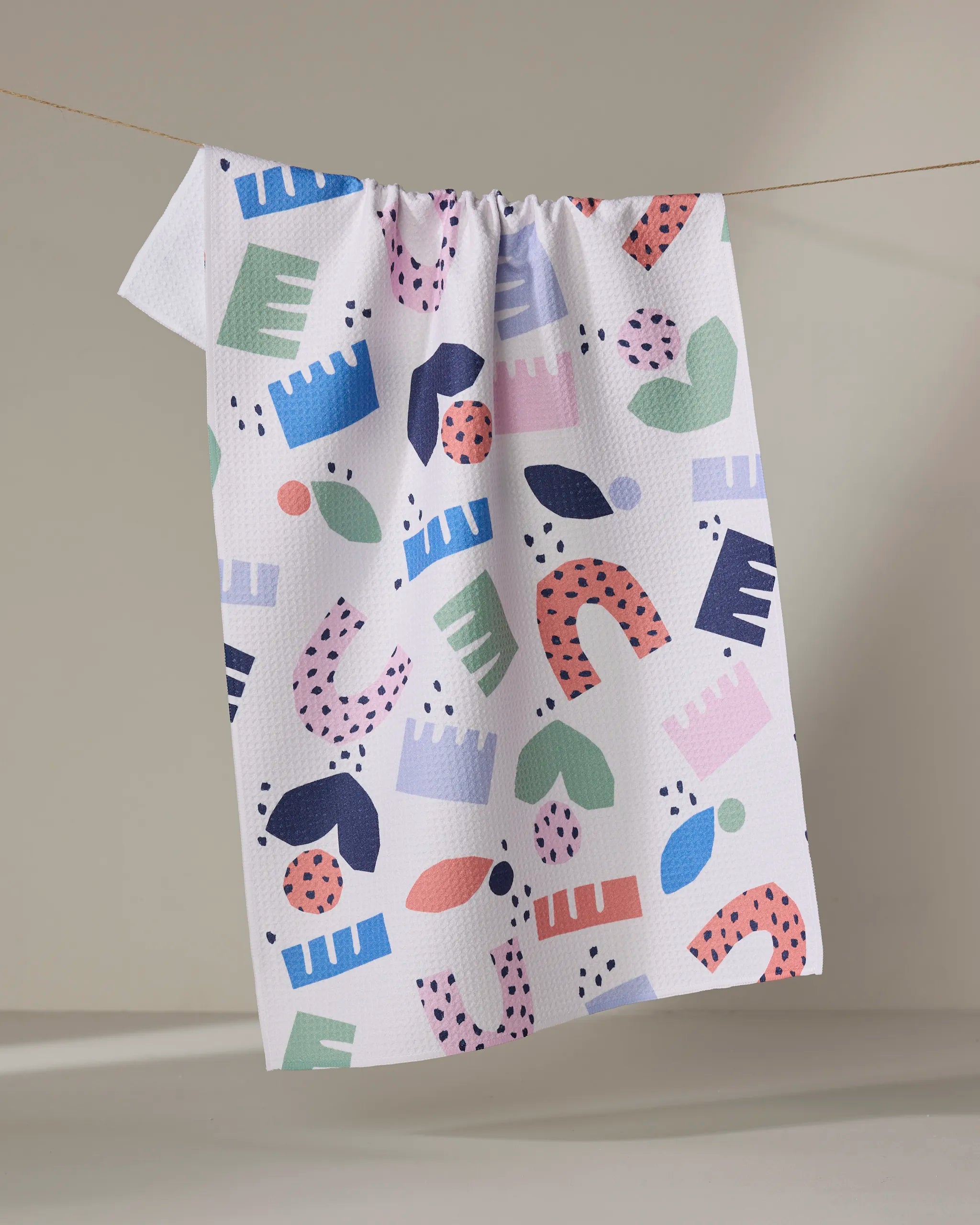 The "Love Letters" tea towel by Claire Iglesias is eco-friendly and white, featuring colorful abstract shapes in blue, green, pink, red, and purple. It hangs on a clothesline against a neutral backdrop.