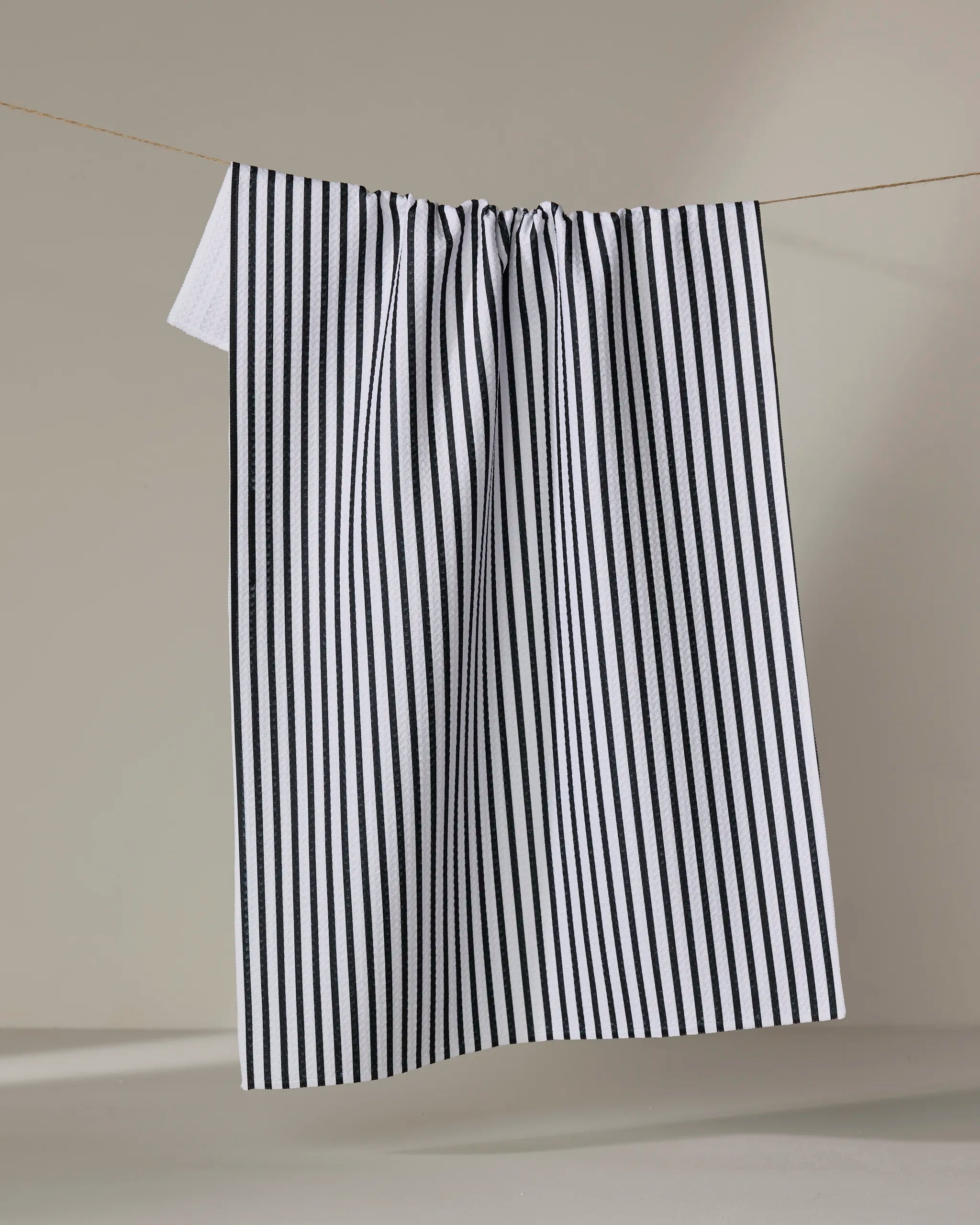 The Kailee Wright Love Notes kitchen towel, featuring black and white vertical stripes and quick-dry fabric, hangs neatly on a clothesline against a simple neutral background.